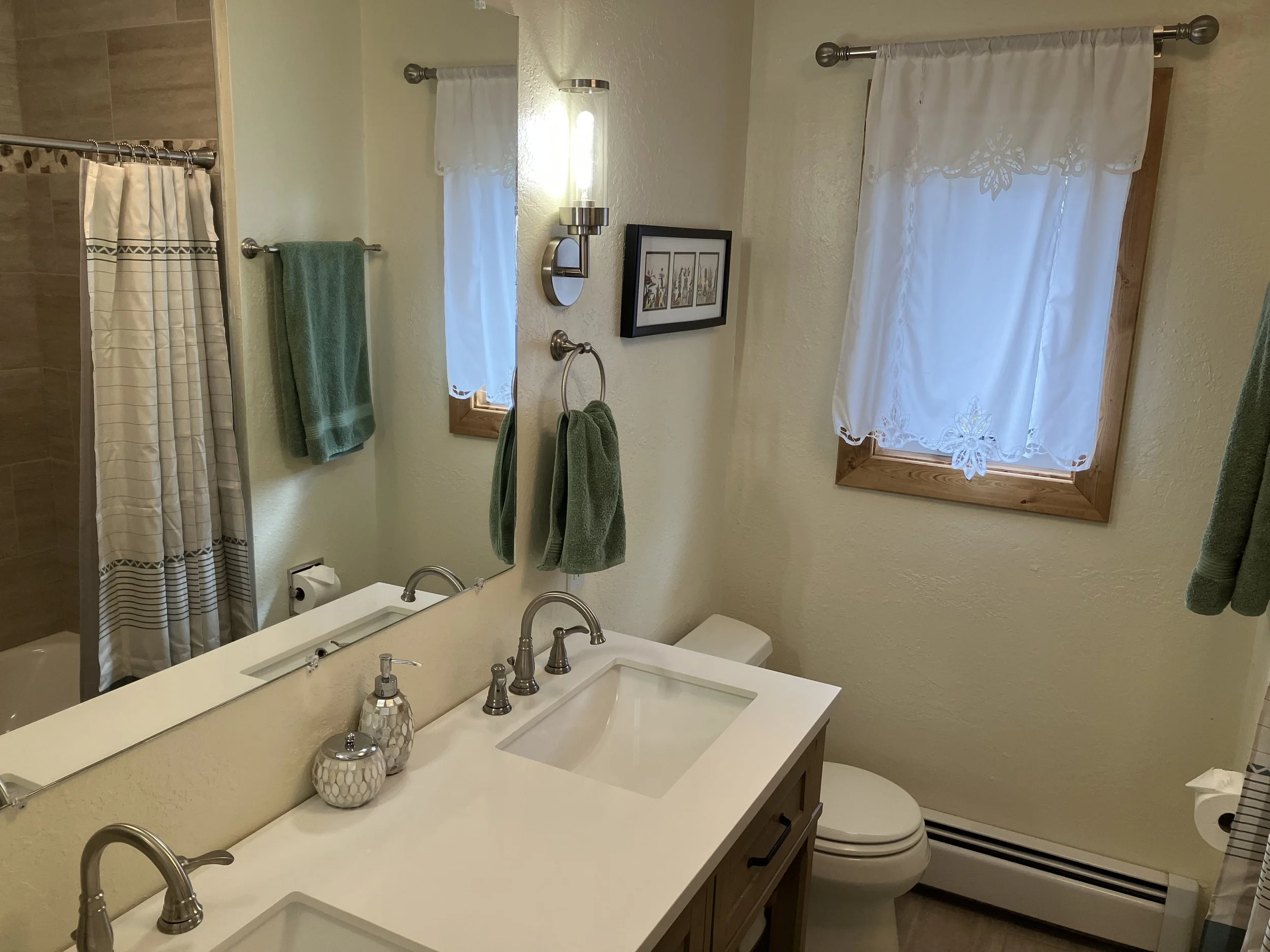 A bathroom with a double sink vanity, large mirror, two small windows with white curtains, green towels, wall-mounted light fixtures, and a shower with a patterned curtain.