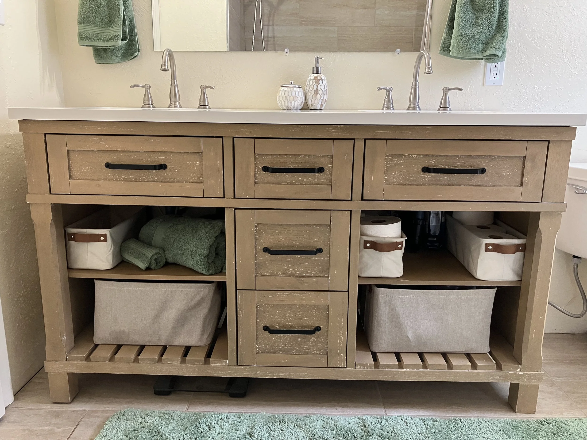 Wooden bathroom vanity with a white countertop, two silver faucets, and candles, with open shelves underneath holding towels and storage bins.