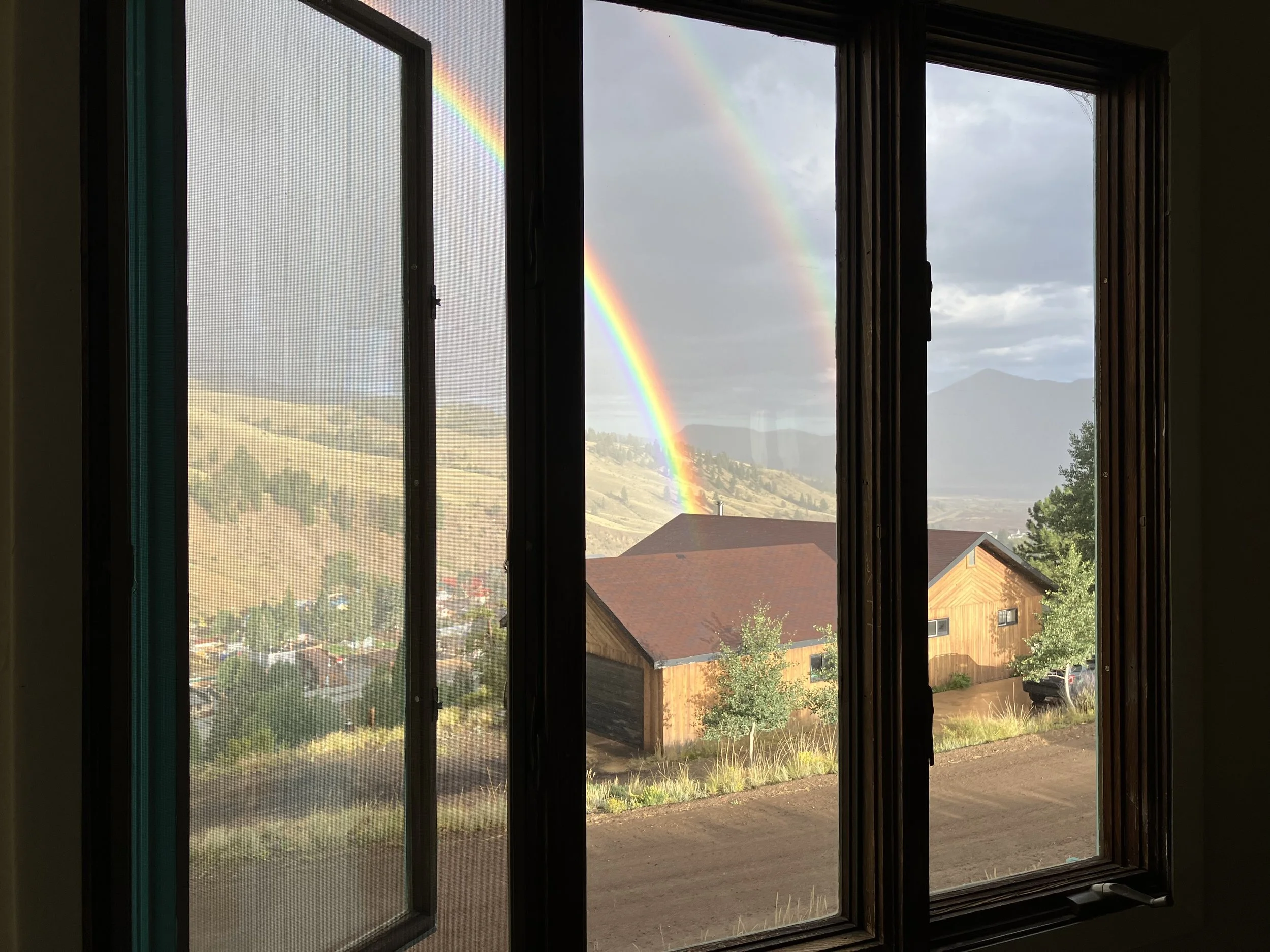 View of a double rainbow outside a window, with hills, houses, trees, and mountains in the background.