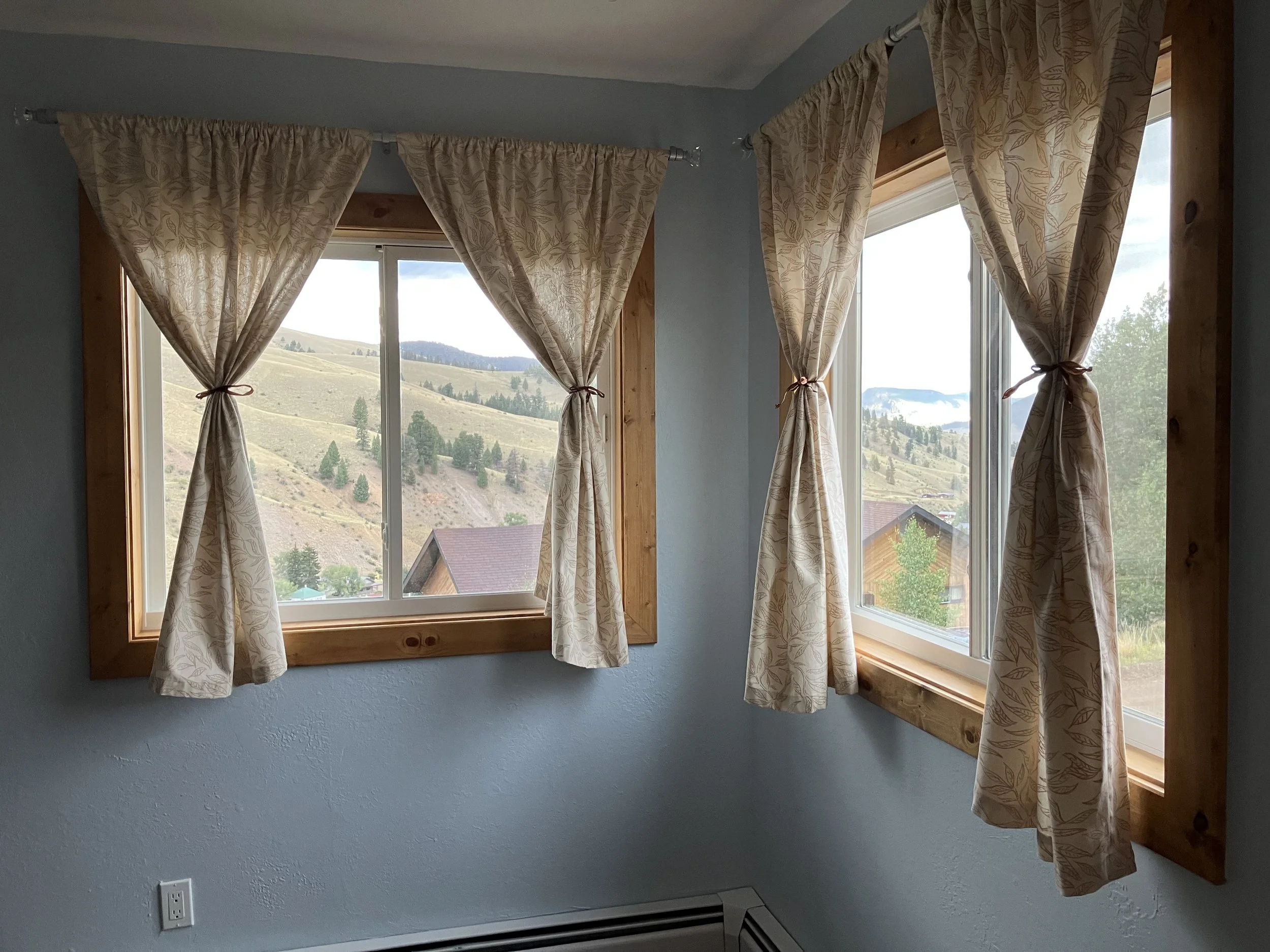 View through two windows with beige, patterned curtains tied back, showing a hilly landscape with trees and houses outside.