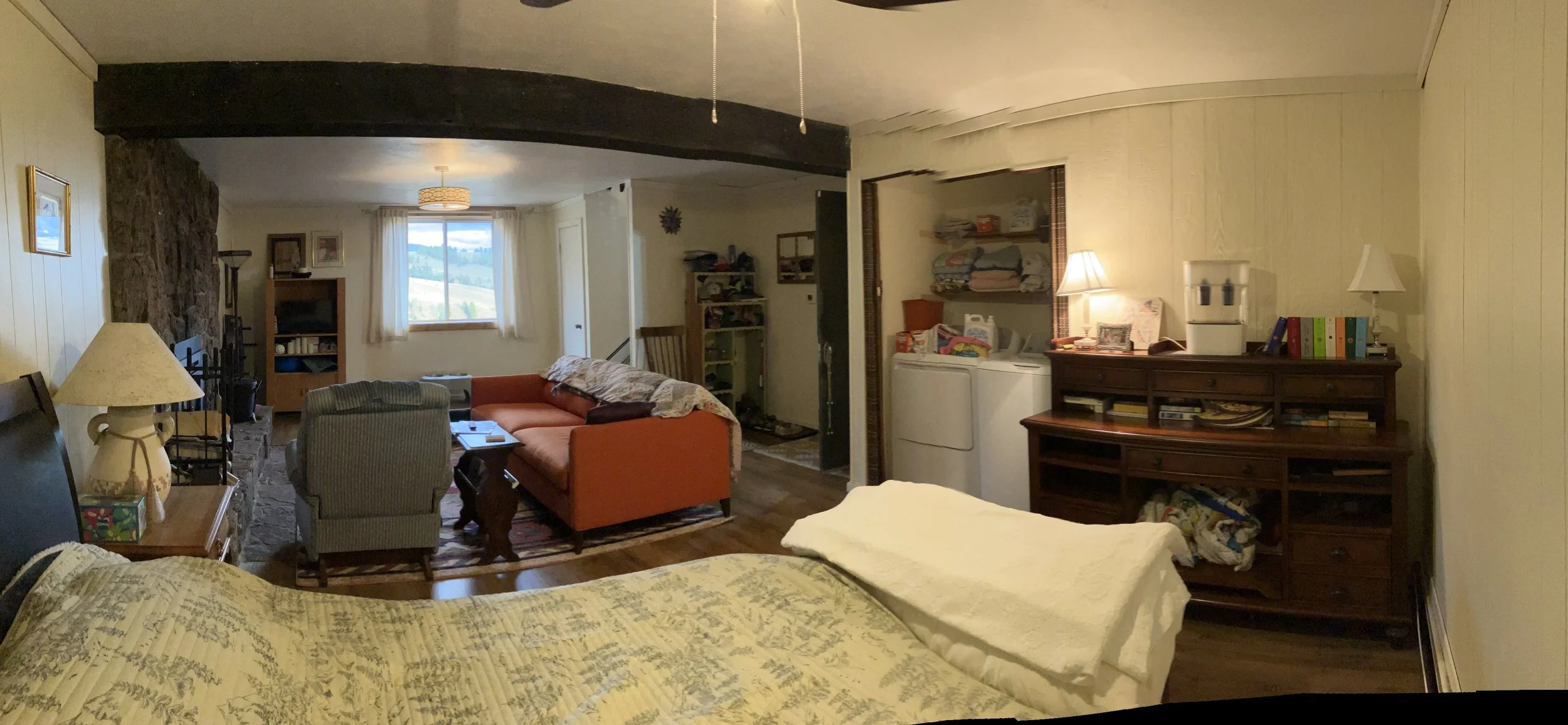 A cozy living room and bedroom with wooden floors, beige walls, and various furniture including beds, couches, a lamp, a bookshelf, and a window with a view of a landscape.