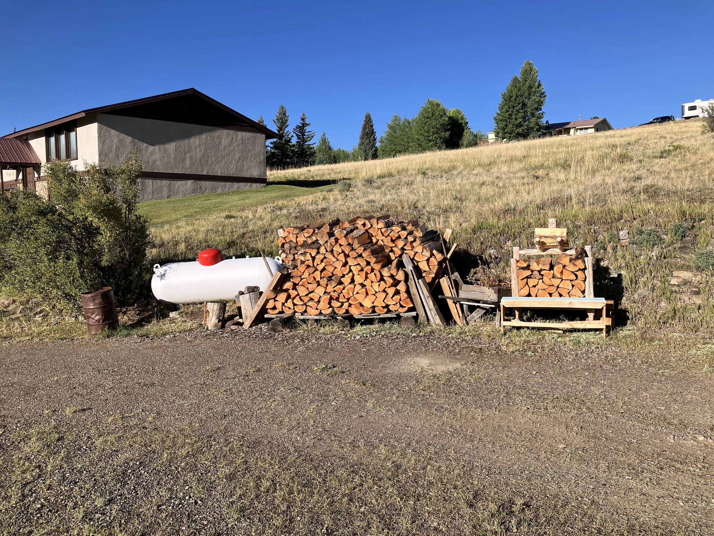 A large pile of cut firewood stacked on a hillside, with a smaller woodpile to the right, a propane tank on the left, and a trash can nearby, under a clear blue sky.
