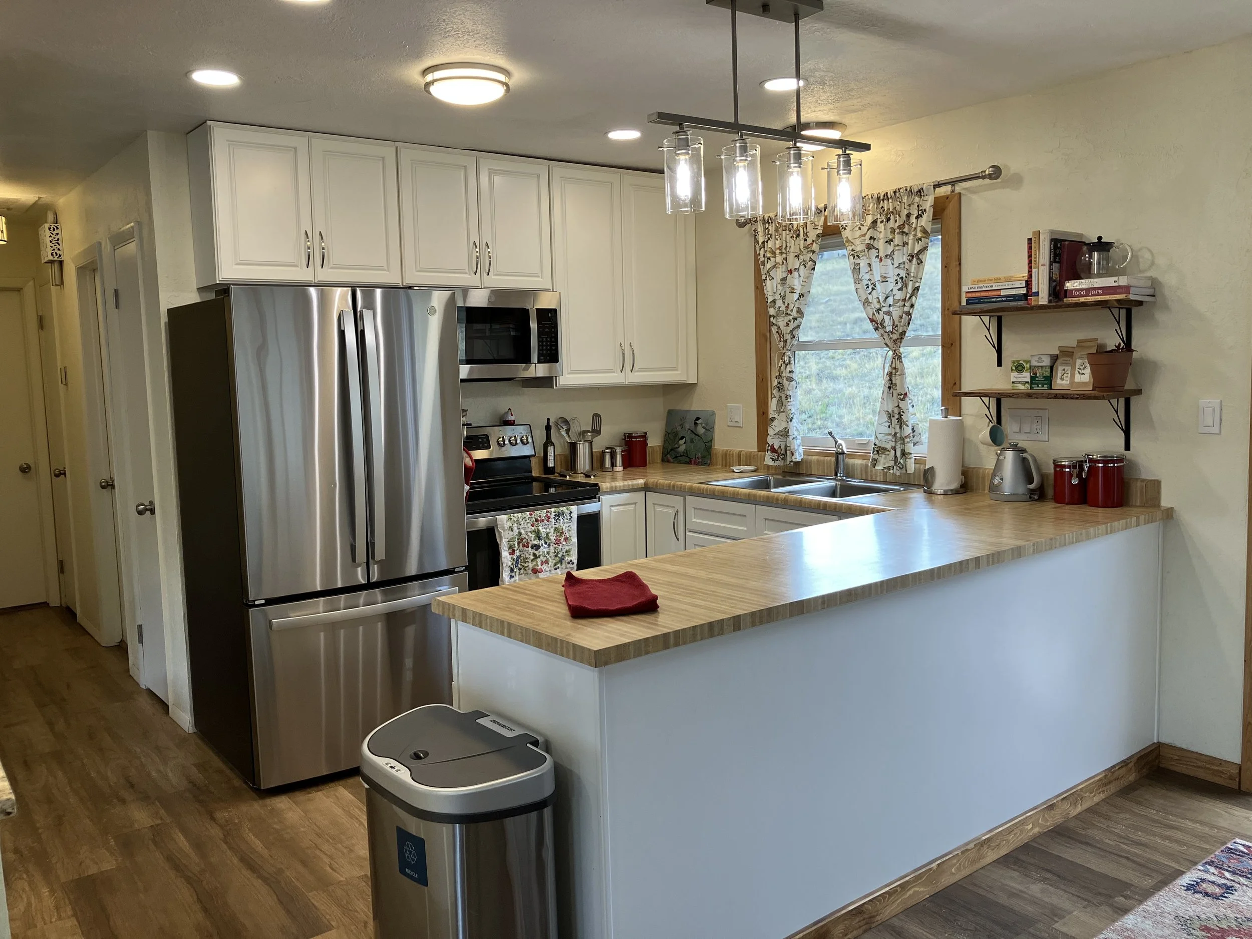 A kitchen with white cabinets, stainless steel refrigerator, microwave, and oven. A wooden countertop with a red dish cloth, a trash can, and blue and red canisters. A window with floral curtains above the sink, with shelves holding books, plants, an