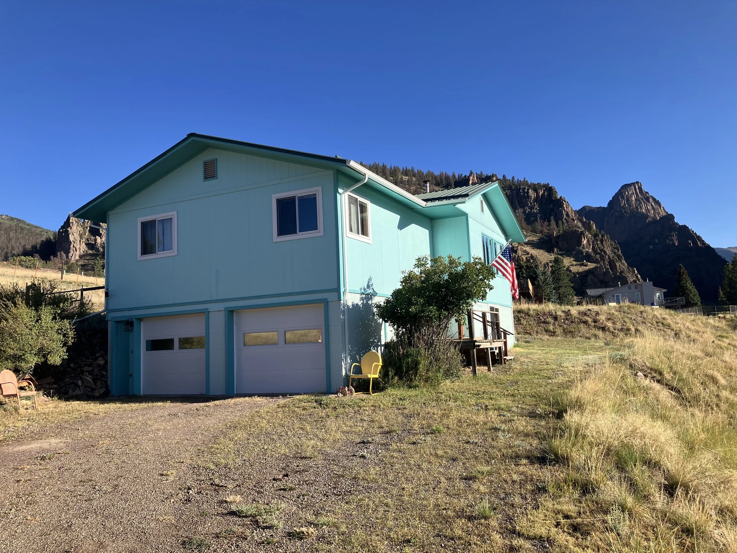 A turquoise two-story house with a garage, set against a mountainous landscape, with an American flag hanging outside, and a small yellow chair next to a tree.