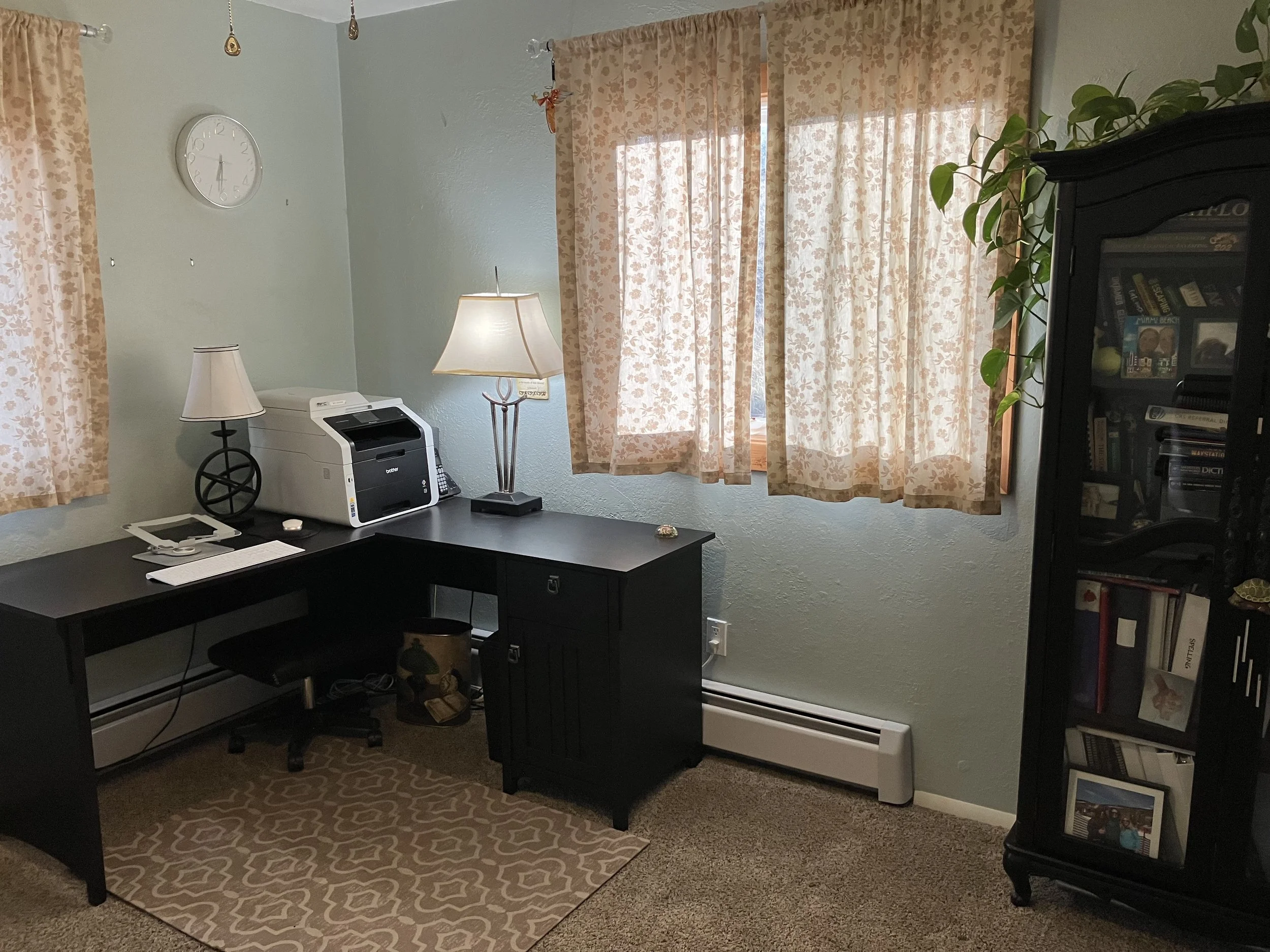 A home office includes a black desk with a printer, a telephone, notebooks, and two lamps with decorative bases. There are beige floral curtains on two windows, a beige patterned rug on tan carpet, a light blue wall with a round clock, a black bookca