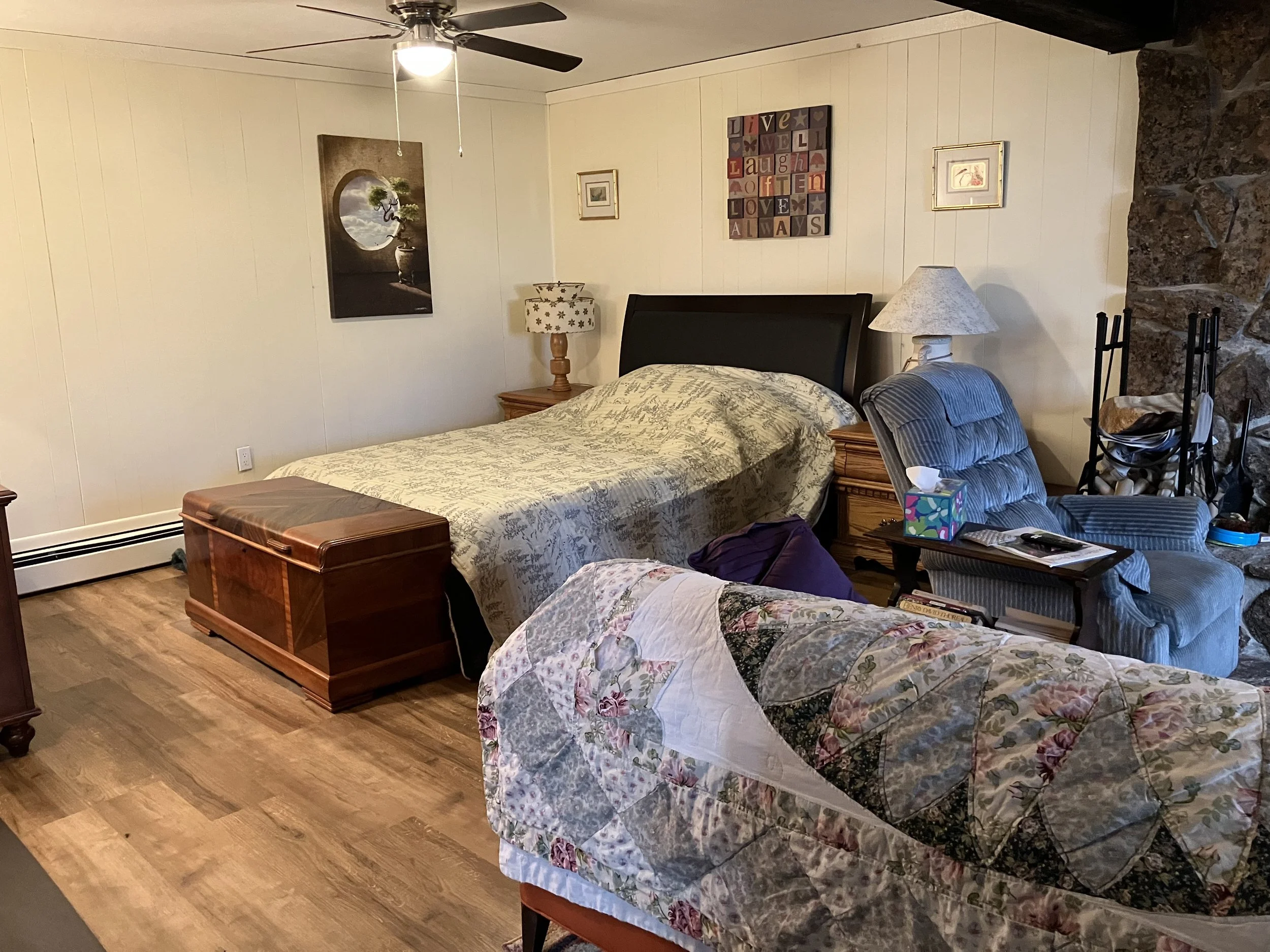 A bedroom with a wood floor, a bed with a beige and green quilt, a wooden chest at the foot of the bed, a rocking chair with a quilt, a blue armchair, and a small wooden nightstand with a lamp. The wall has framed artwork and a decorative sign. A cei
