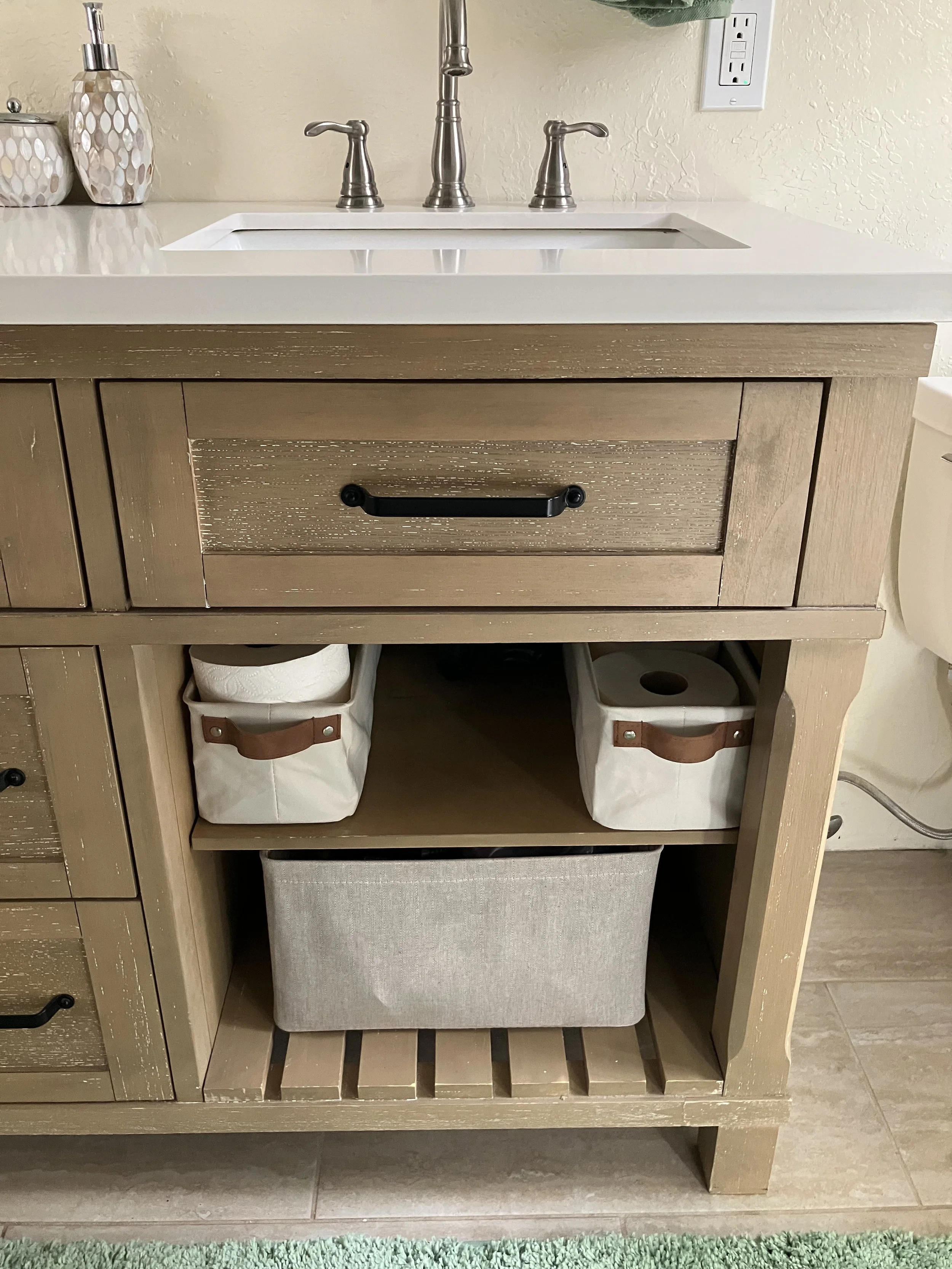 Bathroom vanity with a white countertop and a sink, below it are open shelves containing paper towels, toilet paper, and a fabric storage bin.