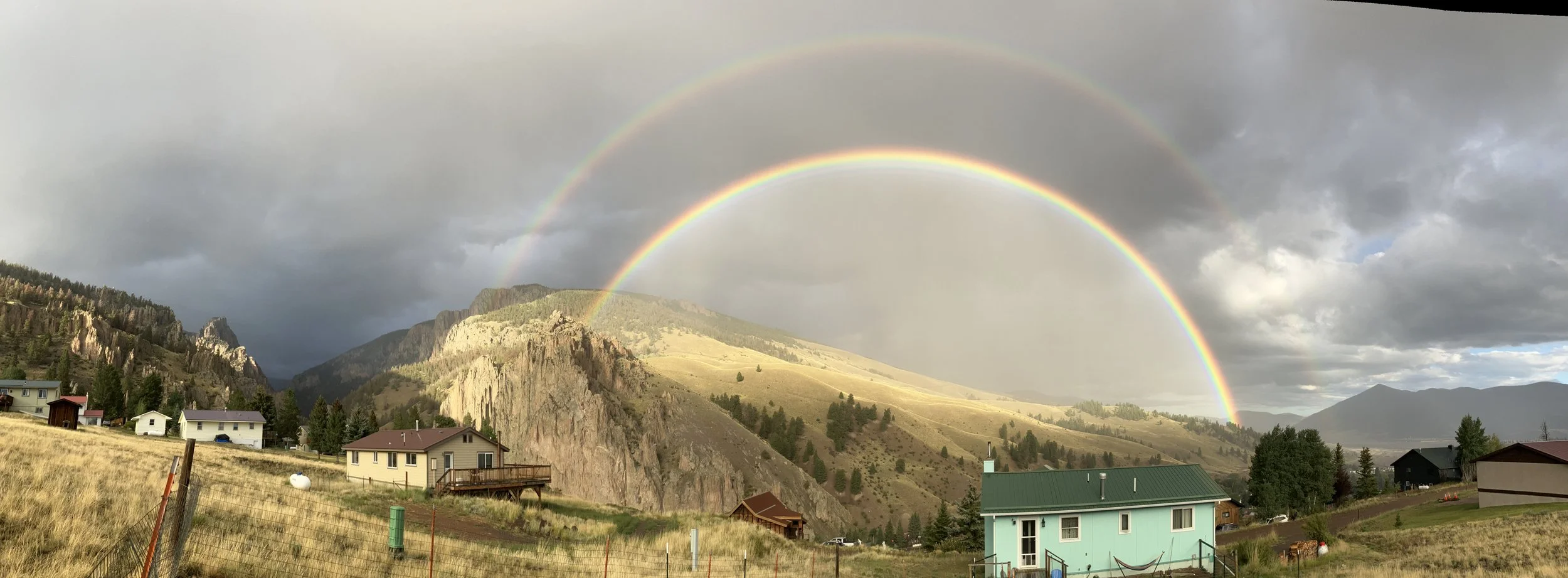 A scenic landscape of houses and rolling hills with a mountain backdrop, under a sky with dark and light clouds, featuring a vibrant rainbow and a faint secondary rainbow.