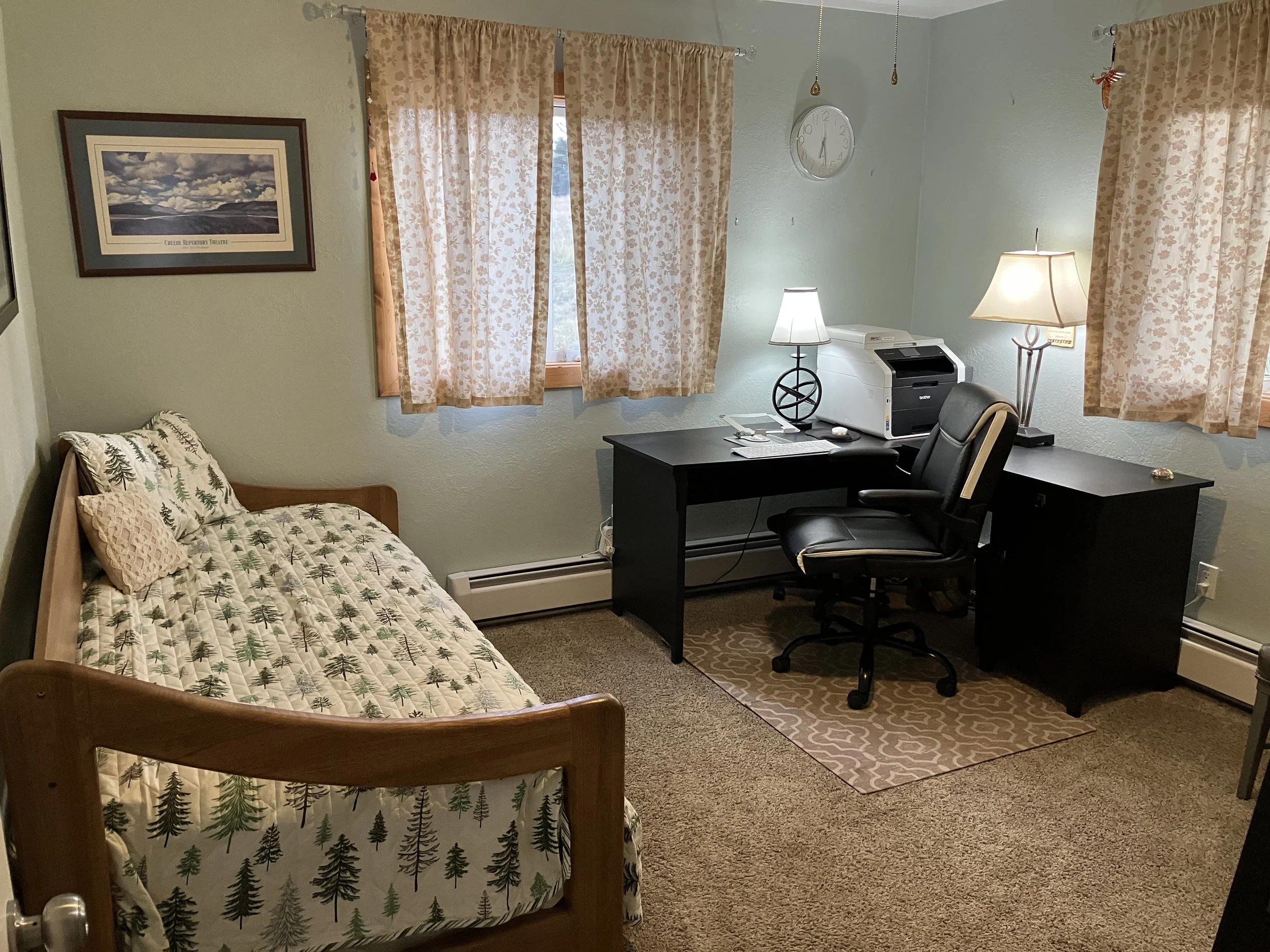 A small bedroom with a single bed and a black desk with a chair. The bed has a pine tree patterned quilt with matching pillows. The room has beige curtains, a clock, and two table lamps. The desk has a printer, phone, and papers.