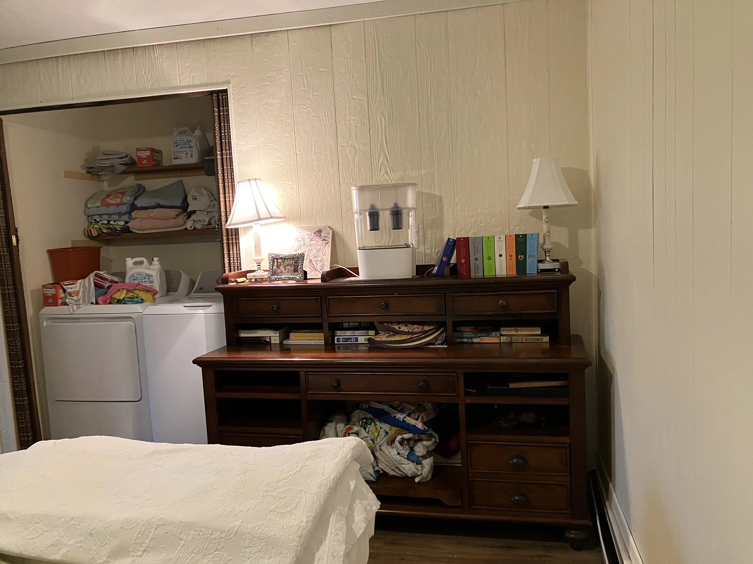 Living room with wooden dresser, table lamp, water filtration system, books, and laundry area with detergents and folded clothes.