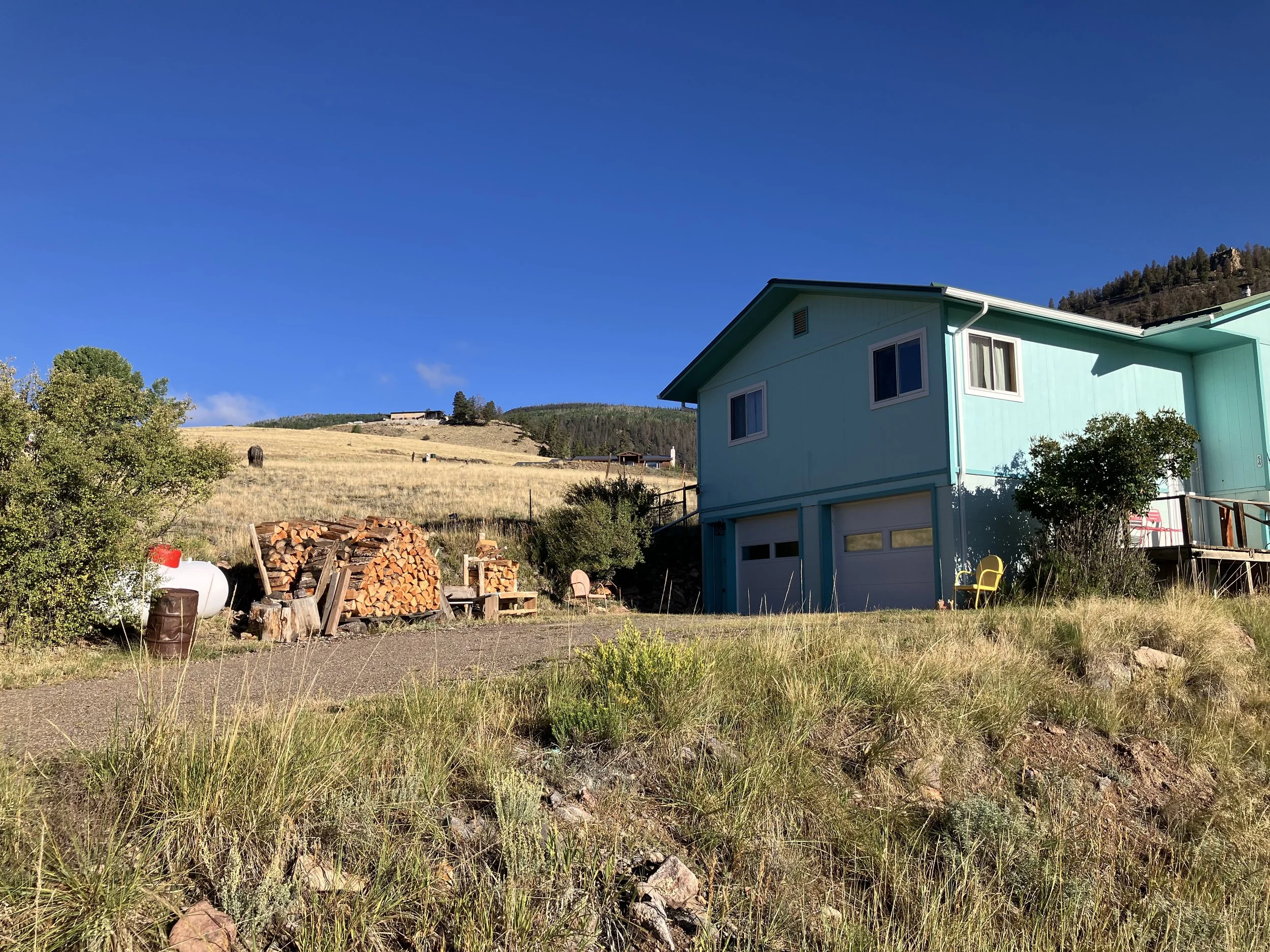 A turquoise house with a white garage door in a rural area, with a woodpile, a propane tank, and a yellow chair outside. The background features a grassy hillside under a clear blue sky.