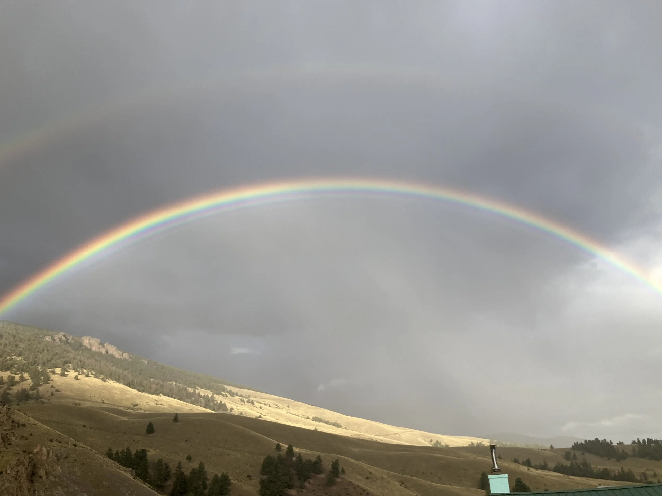 Double rainbow arcing over a hilly landscape with trees, cloudy sky, and a small building at the bottom right.