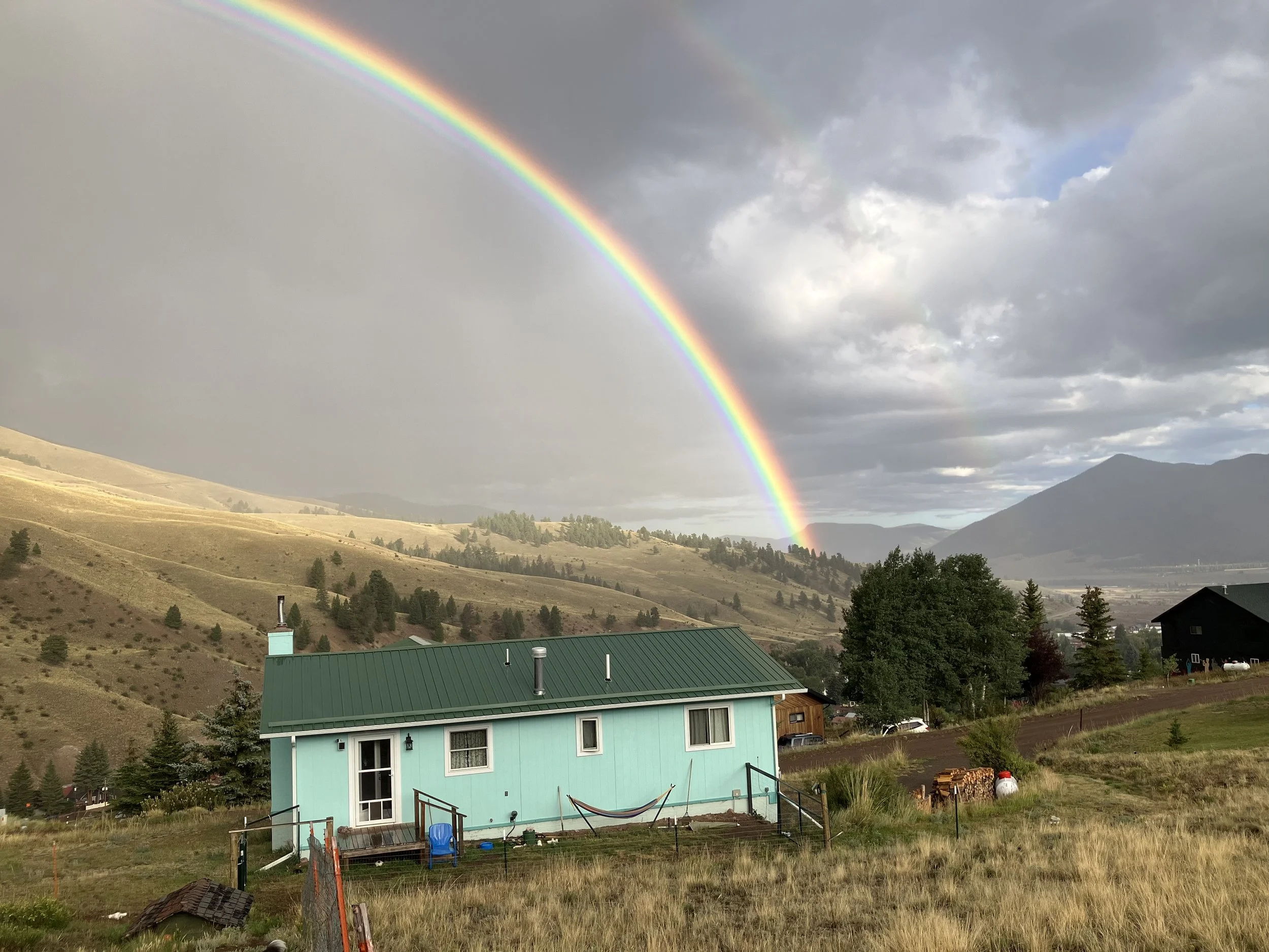 A landscape with a rainbow arching over a green house, rolling hills, and mountains in the background under a cloudy sky.