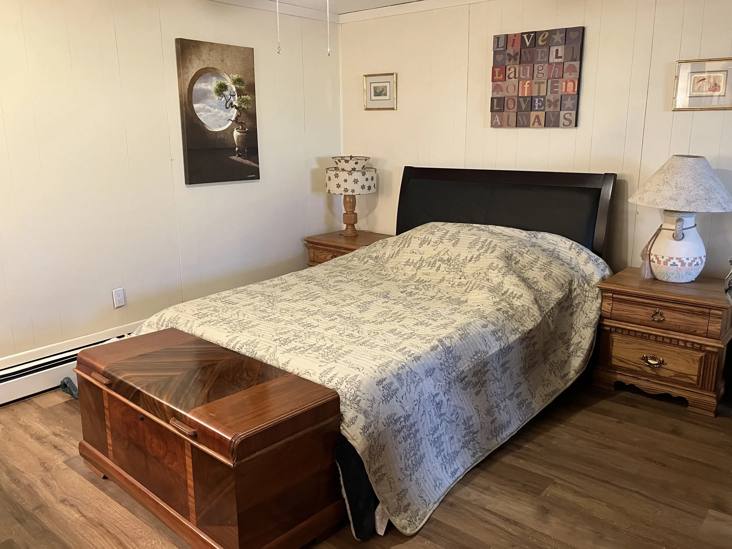 A bedroom with a wooden chest at the foot of the bed, which has a dark headboard and a patterned bedspread. There are two wooden nightstands with lamps, wall art, and a decorative sign above the bed.