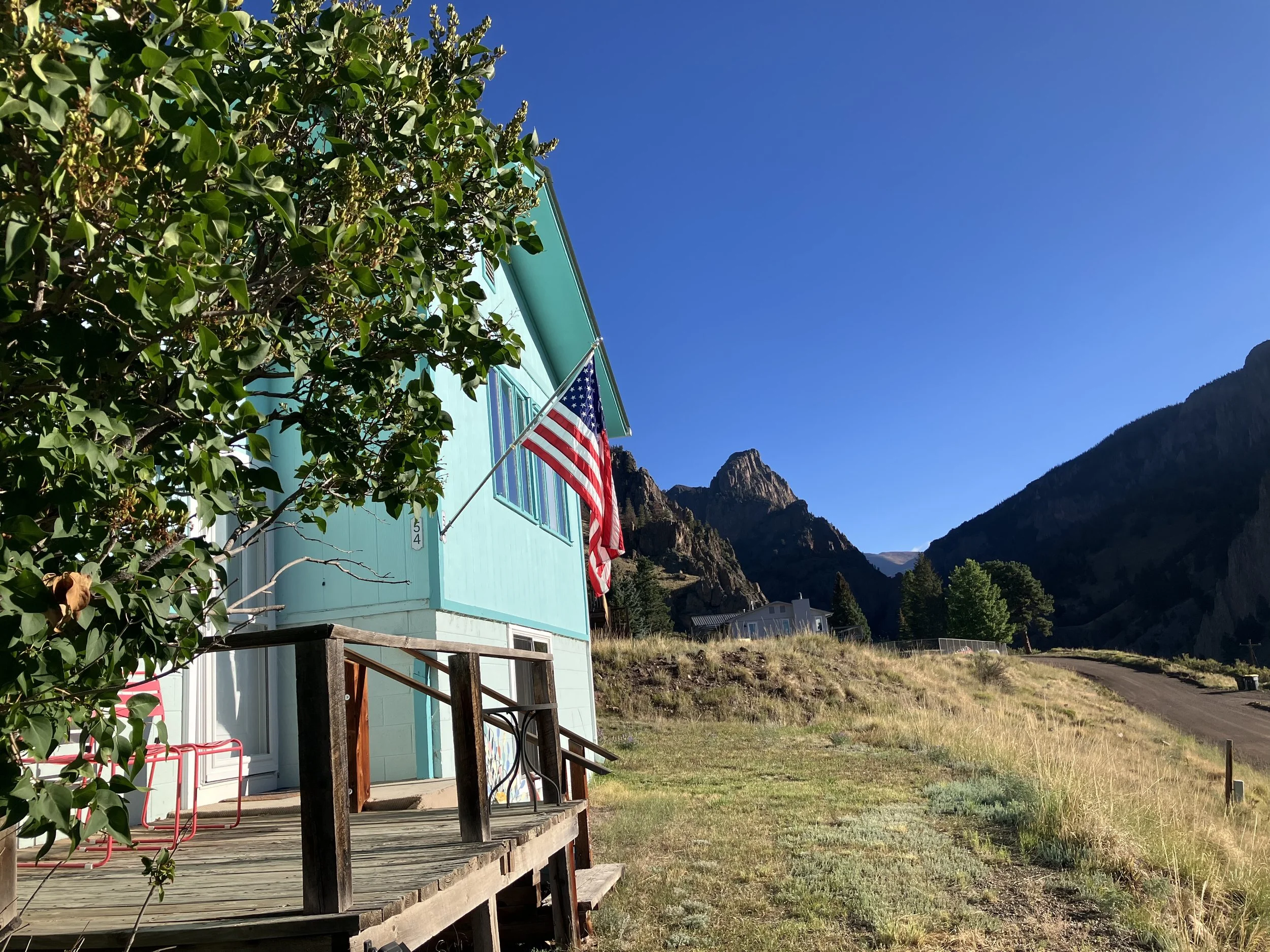 A turquoise house with an American flag hanging from a pole on the porch. The house is on a grassy hillside with a wooden deck. Behind, there are rocky mountains and a clear blue sky.