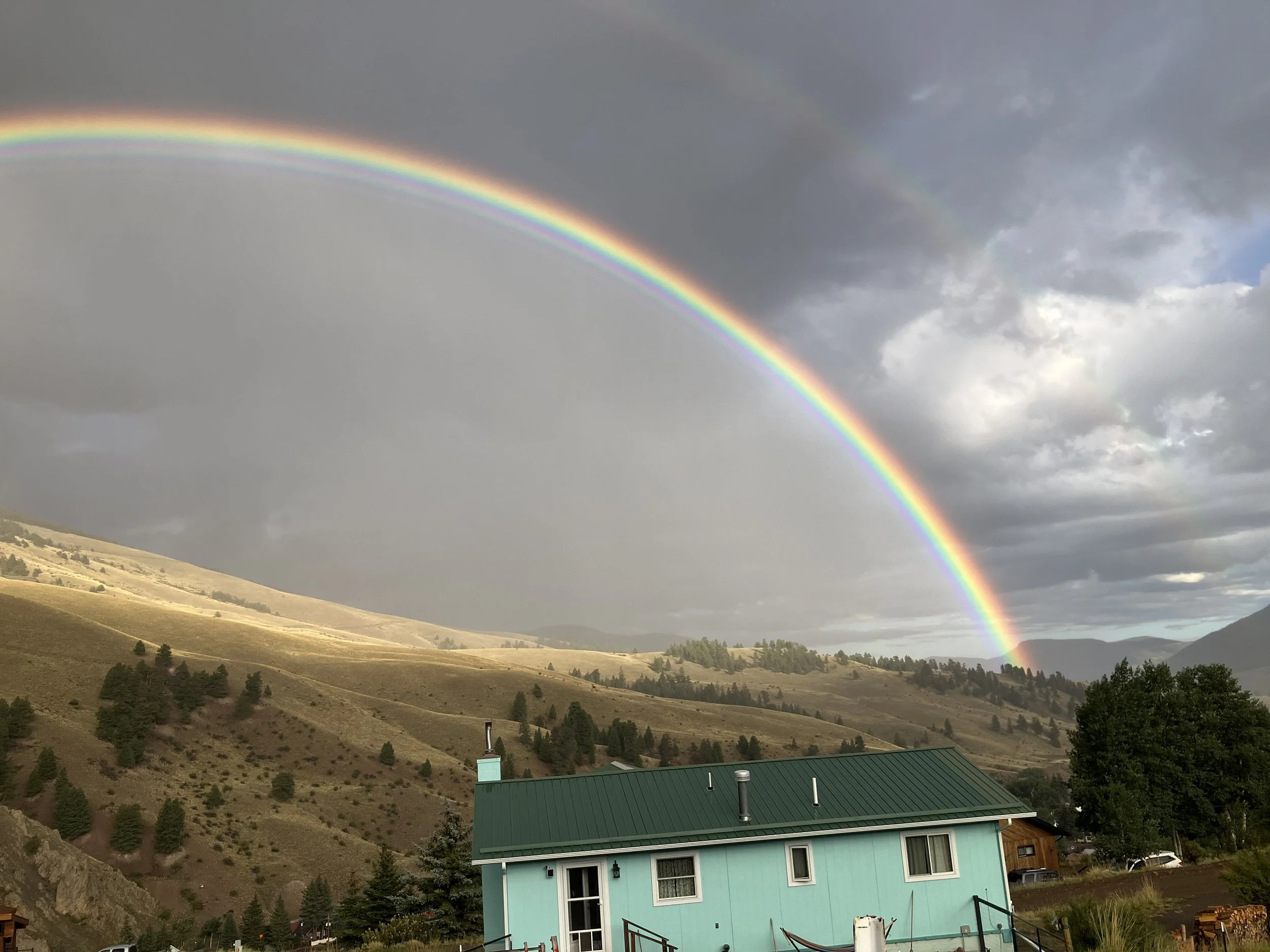 Rainbow arching over a green house with mountains and cloudy sky in the background.