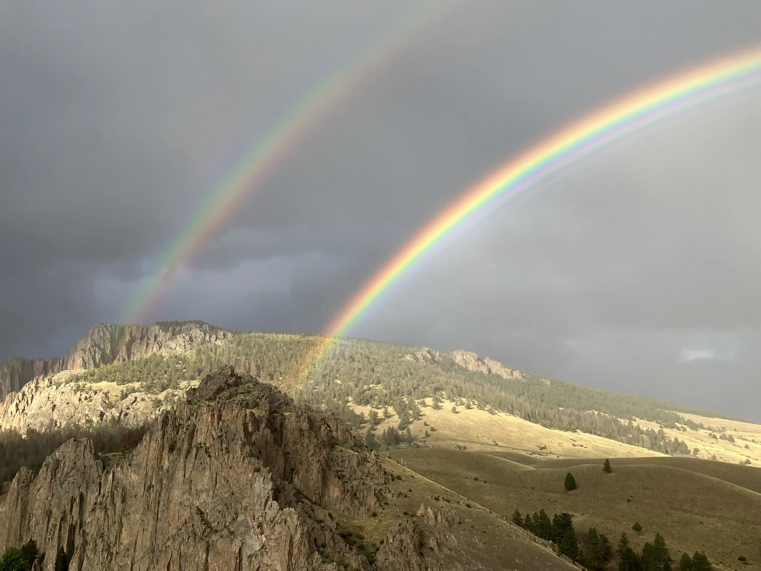 A scenic view of two rainbows arching over a rugged mountain landscape, with cloudy skies overhead.