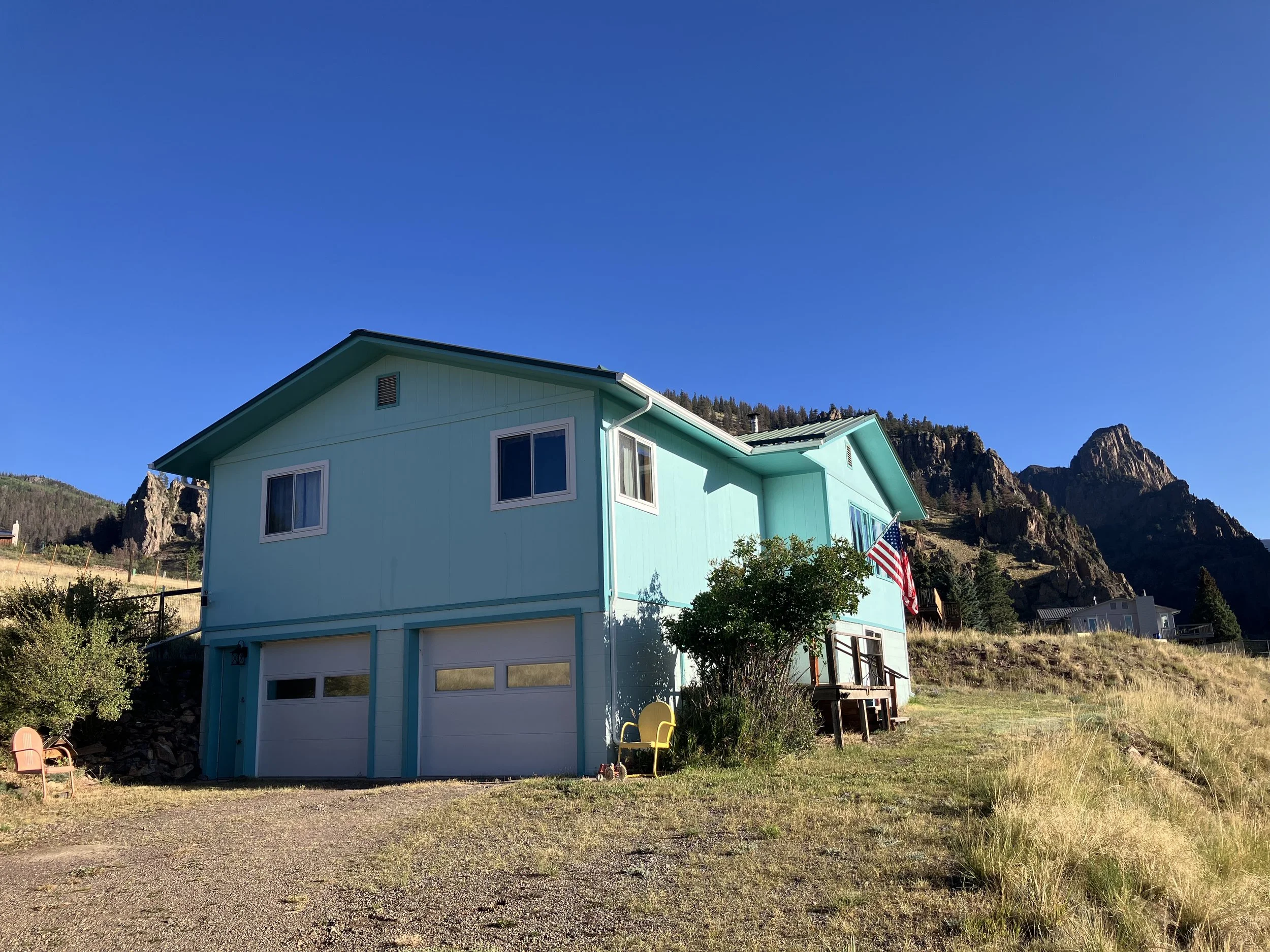 A light blue house with a garage door, set against a backdrop of rocky mountains and clear blue sky.