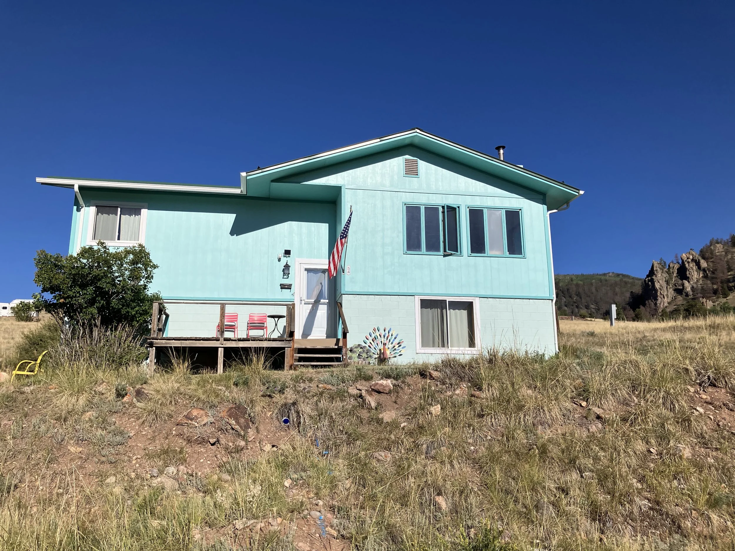 A sky-blue two-story house with four large windows on the upper floor, a small front porch with two chairs, and an American flag hanging beside the door, set against a clear blue sky and a grassy hillside.