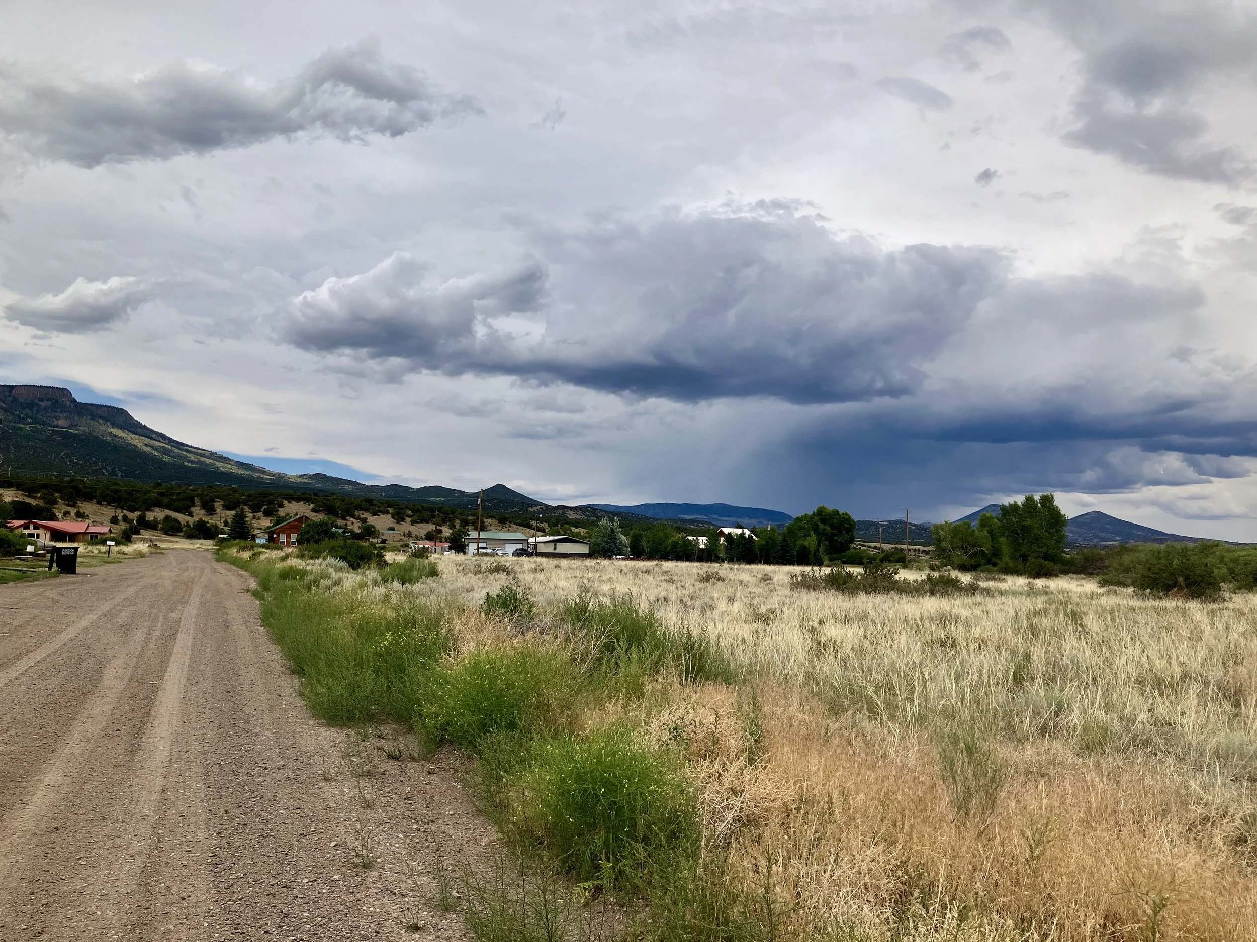 A dirt road running alongside a grassy field with houses and trees, mountains in the background, under a cloudy sky with dark storm clouds.