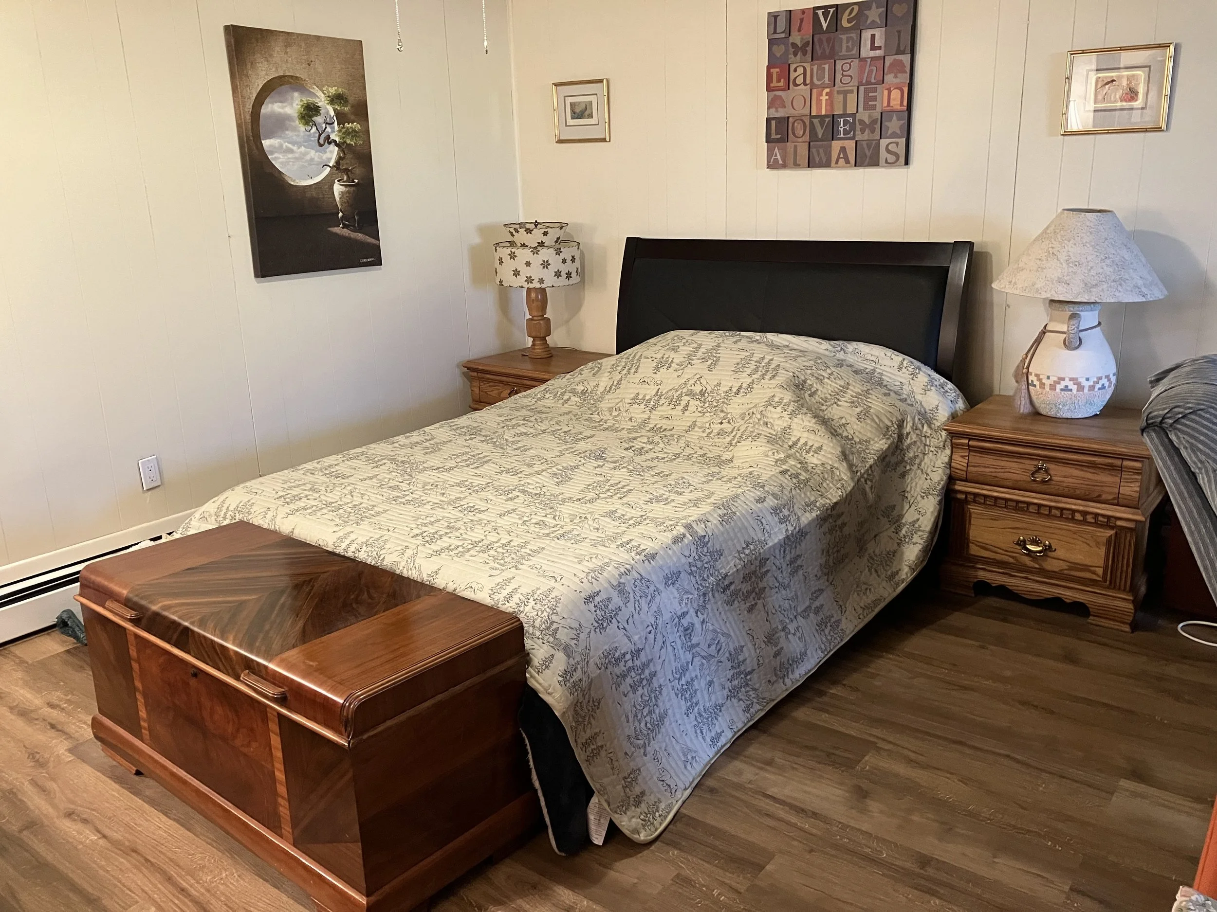 A bedroom with a bed, two wooden nightstands with lamps, and wall art. The bed has a quilt with a toile pattern, and a wooden chest at the foot of the bed. The wall art includes a framed picture, a colorful block design, and botanical prints.
