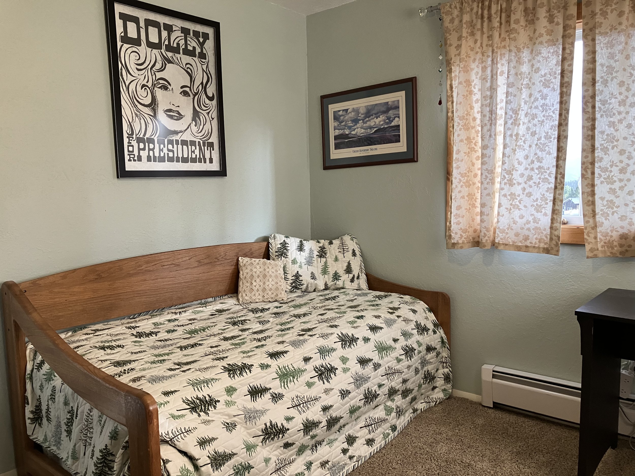 A small bedroom with a wooden daybed covered with a pine tree patterned quilt and two pillows, one plain beige and the other with pine tree designs. The room has a green wall with framed artwork and a large poster of a woman labeled "Dolly for Presid