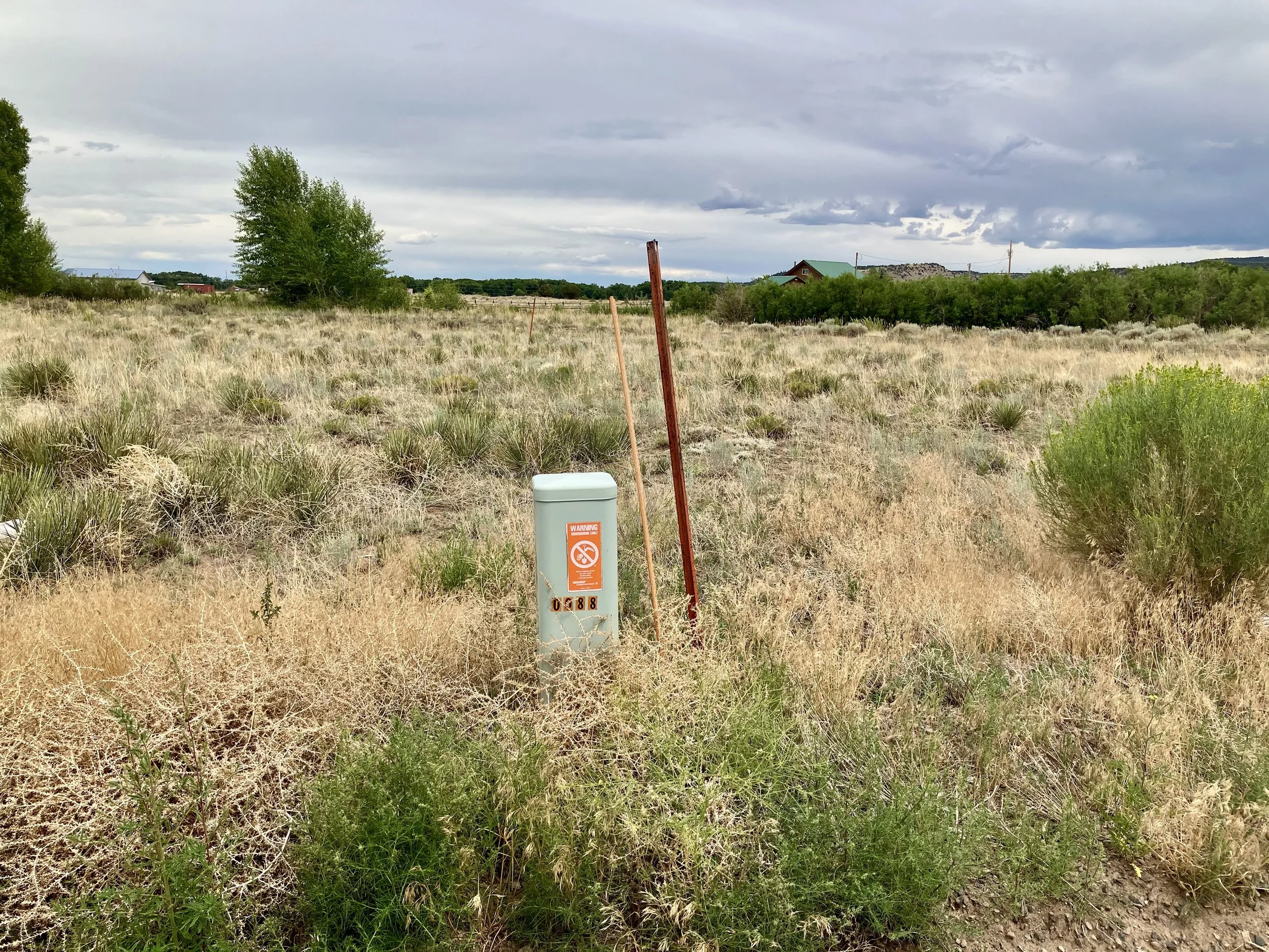 Open field with dry grass and sparse green bushes, a warning sign, and utility poles under a cloudy sky.