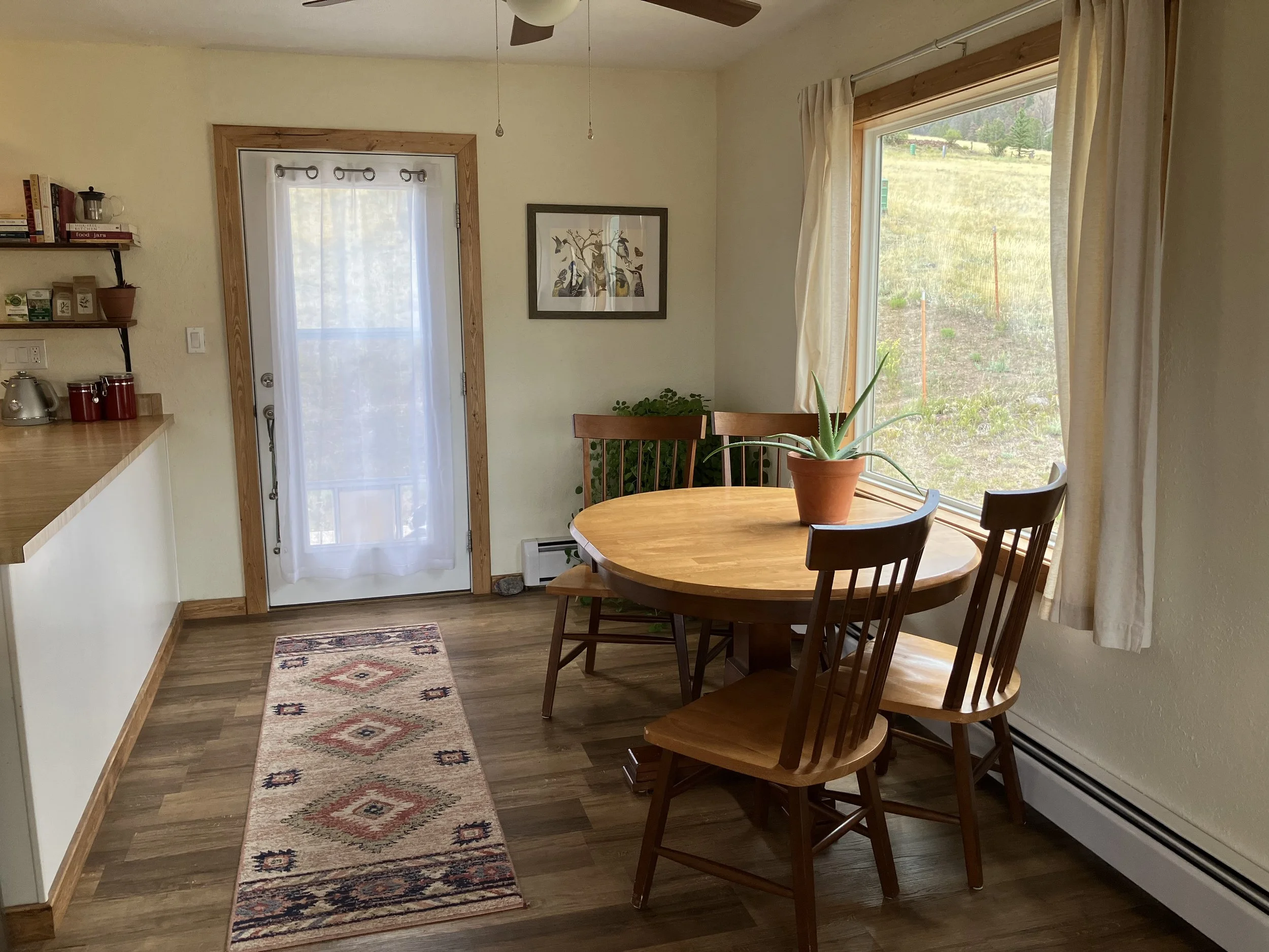 A cozy dining area with a round wooden table and four wooden chairs, one with a potted plant on top, beside a large window with cream curtains. A door with a sheer curtain leads outside, with a view of a grassy yard. The walls are light-colored, with