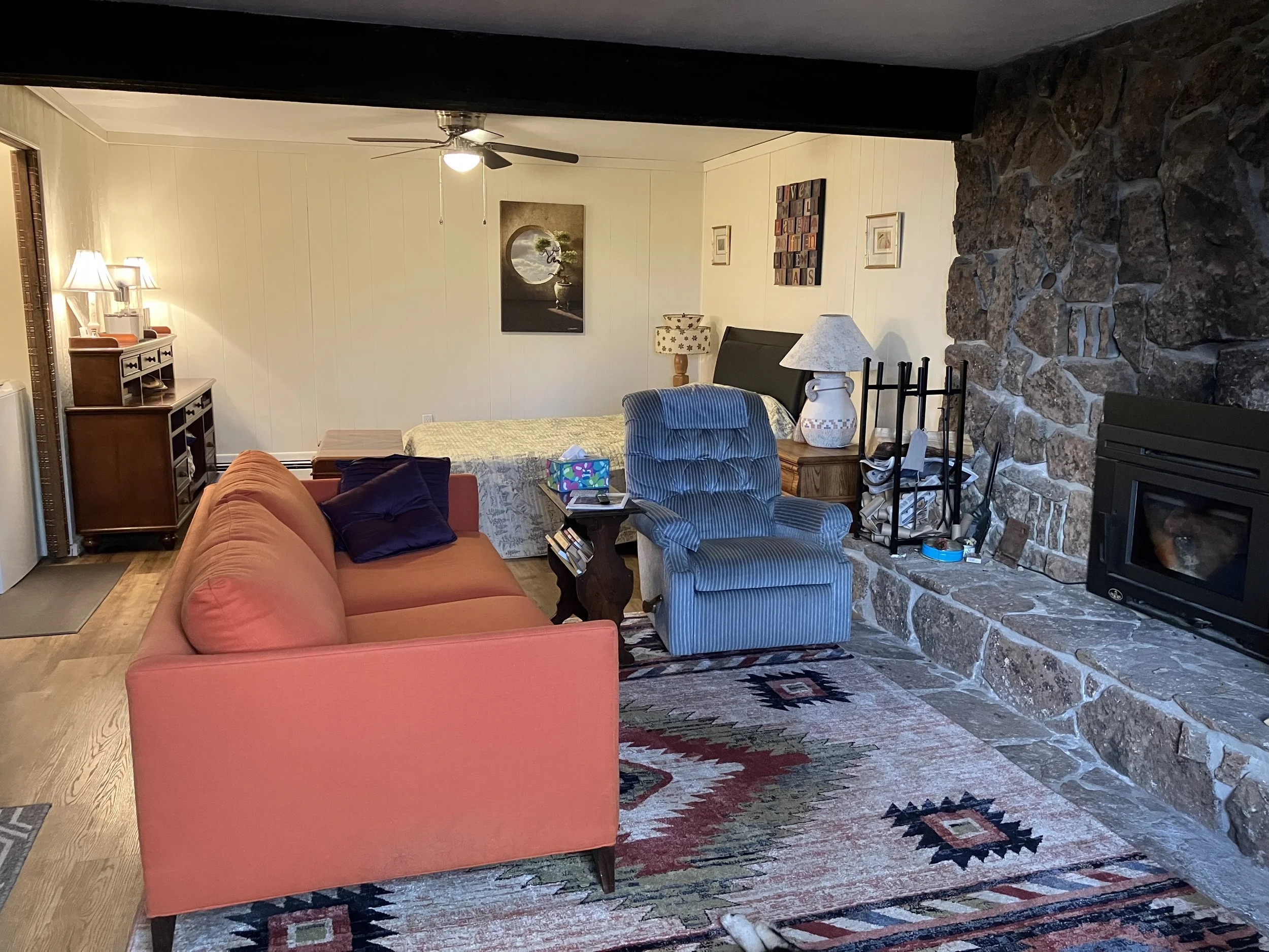 Living room with coral sofa, blue recliner, stone fireplace, wooden side tables, table lamps, and a patterned rug.