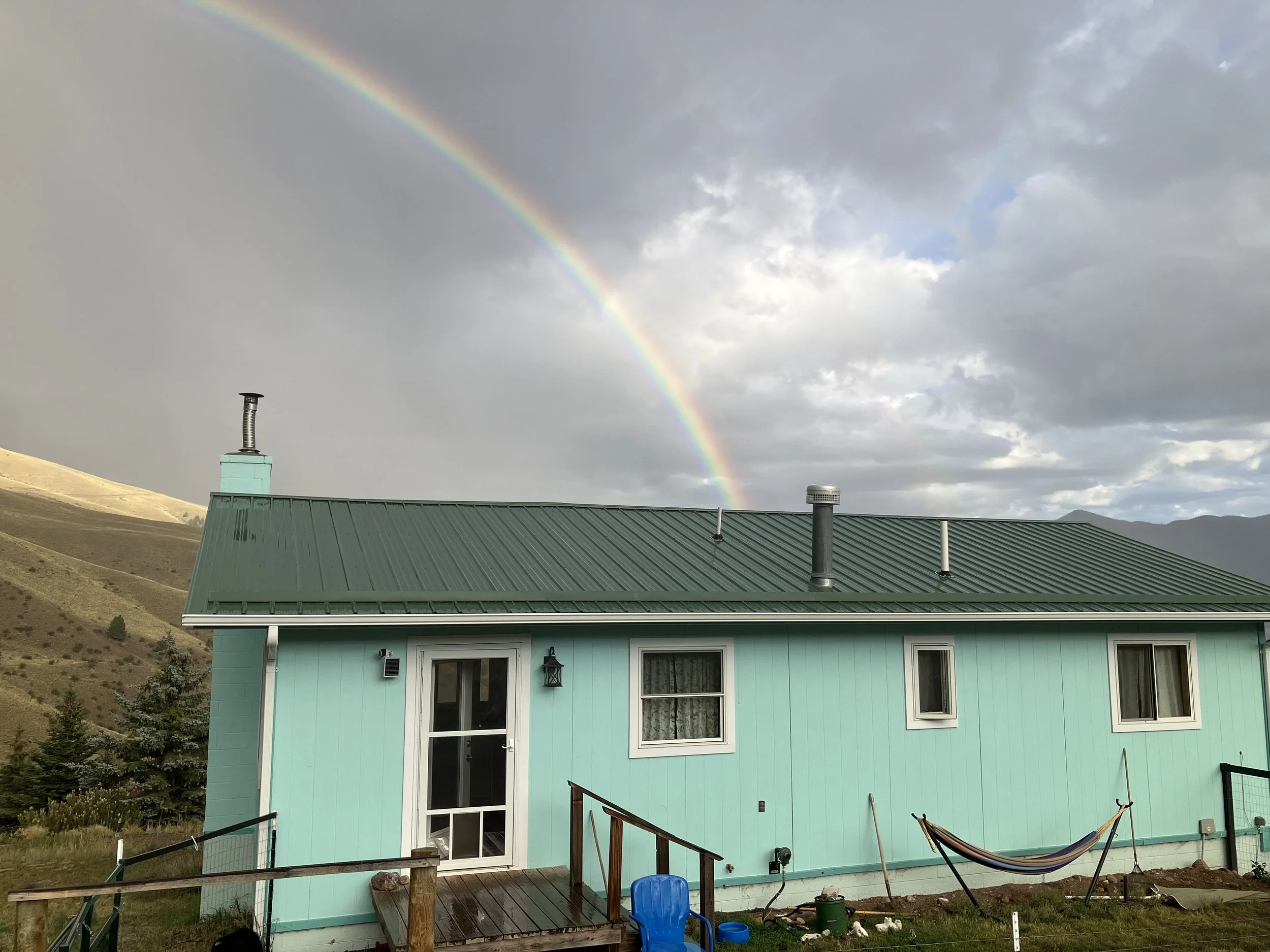 A turquoise house with a green metal roof, three windows, and a door, is set against a backdrop of mountains and a cloudy sky. A rainbow arches across the sky.