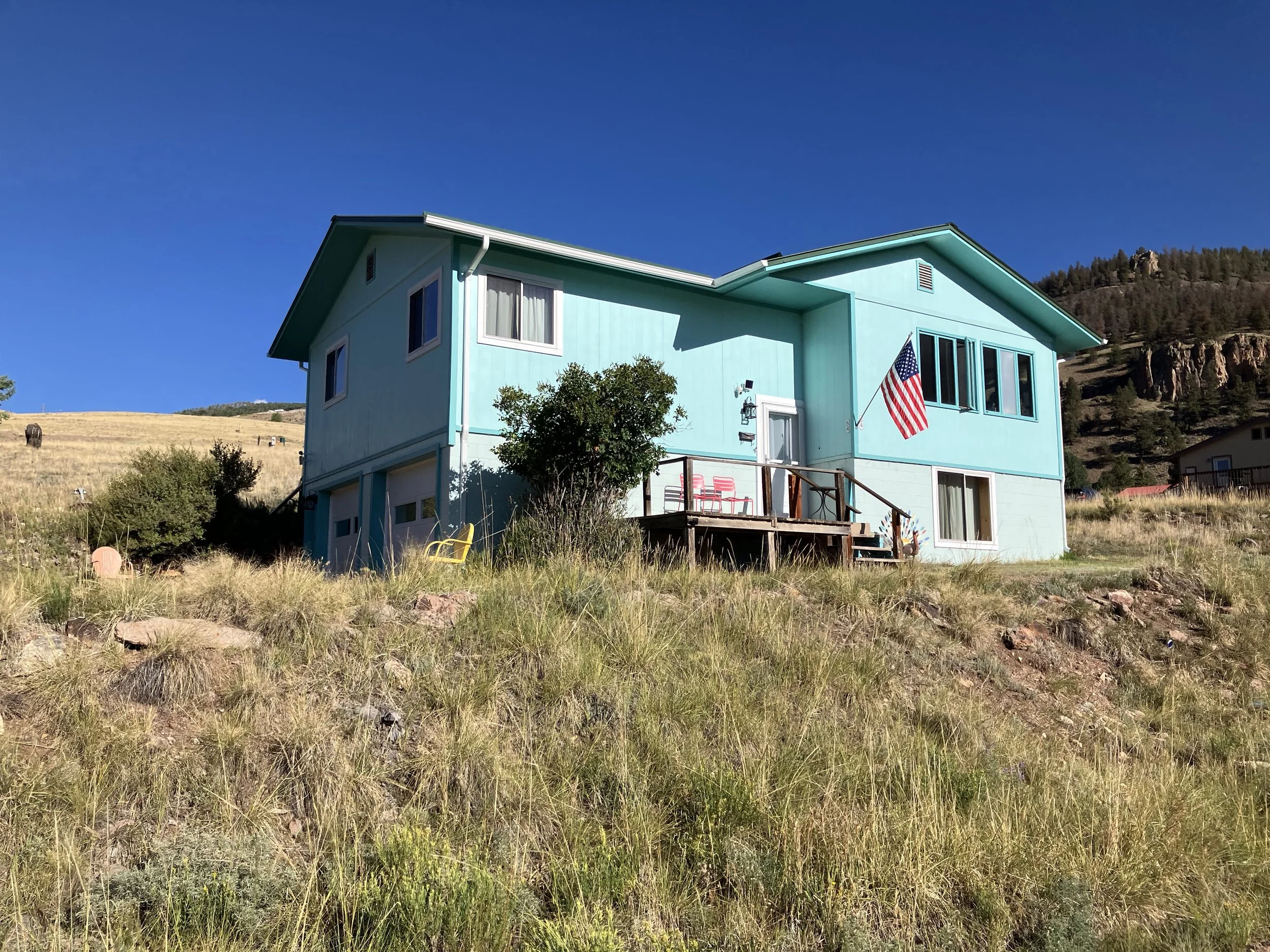 A light blue two-story house on a grassy hillside with a small porch and an American flag. Clear blue sky and some distant hills are in the background.