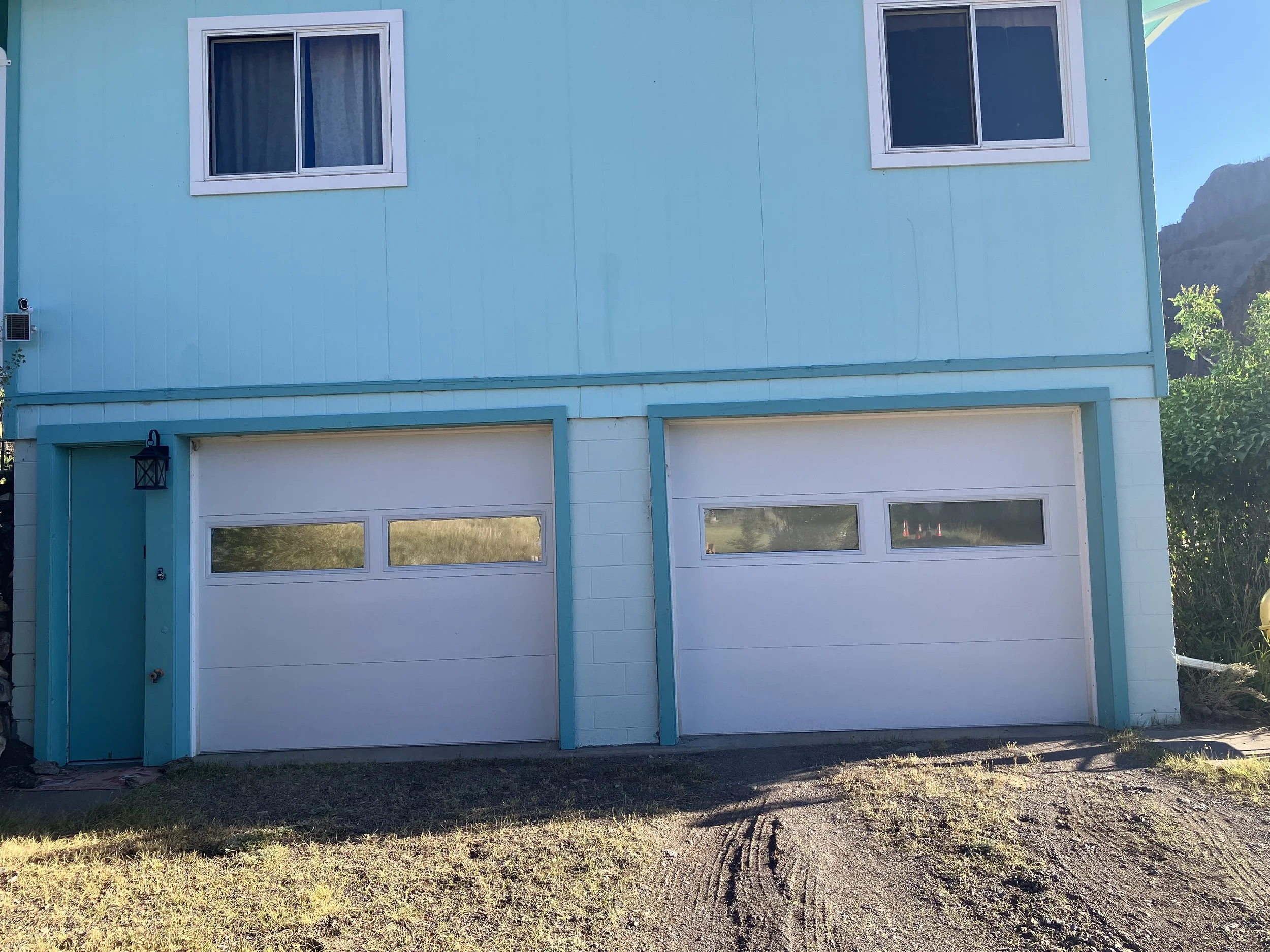 A two-story house with light blue exterior walls, two white garage doors with small rectangular windows, and two windows on the upper level. The house has a small black lantern-style light fixture beside the left garage door. The ground in front is a
