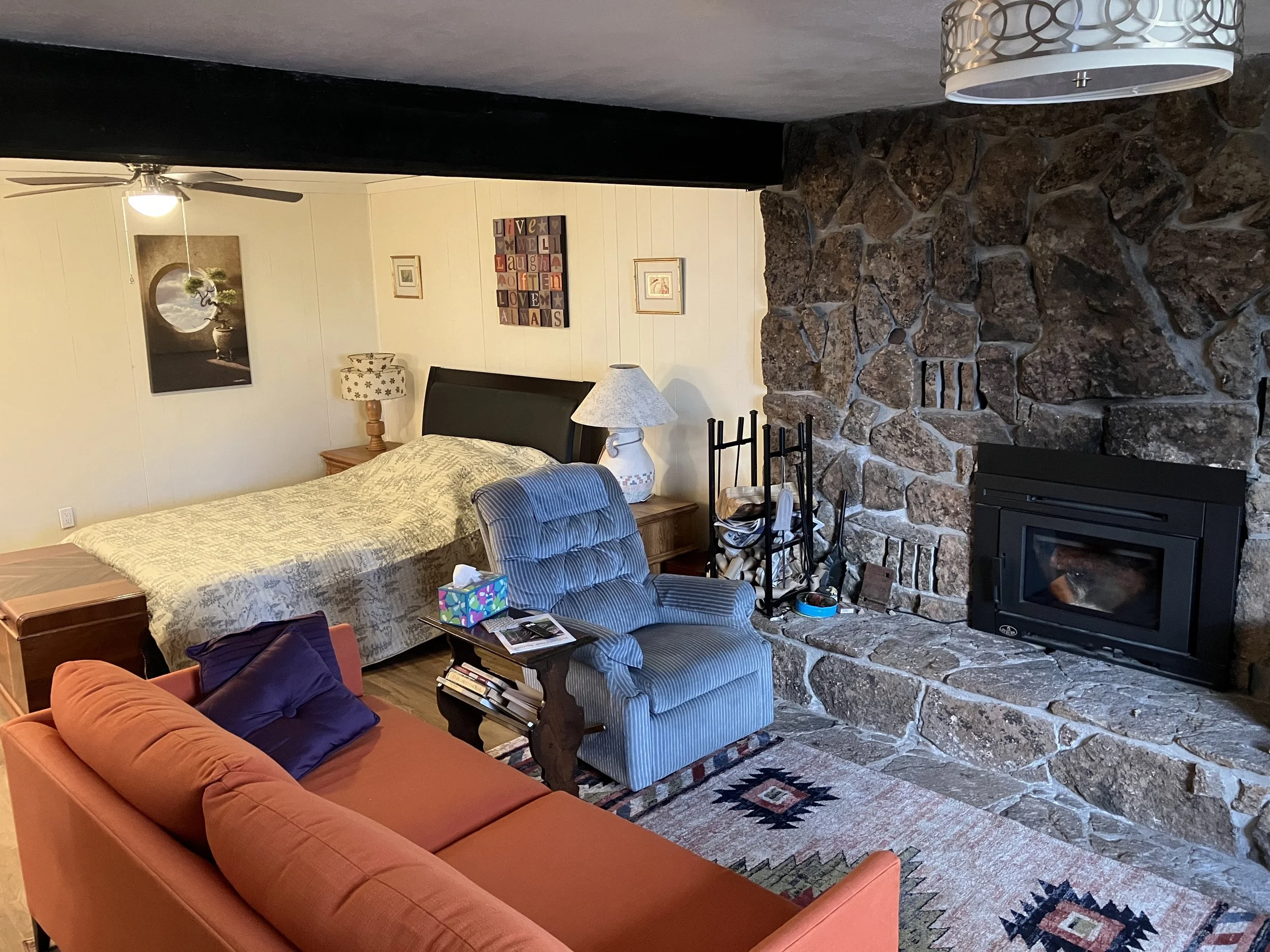 Living room with a stone fireplace, a bed with lamps and artwork on the wall, a reading chair, a couch with pillows, and a rug.