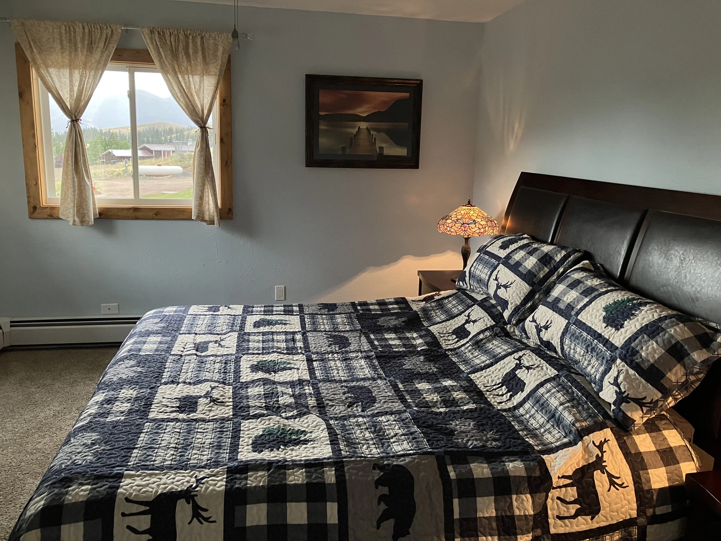 A bedroom with a bed covered in a quilt with moose and pine tree patterns, a nightstand with a stained-glass lamp, a window with cream curtains showing a rural landscape, and a framed landscape picture on the wall.