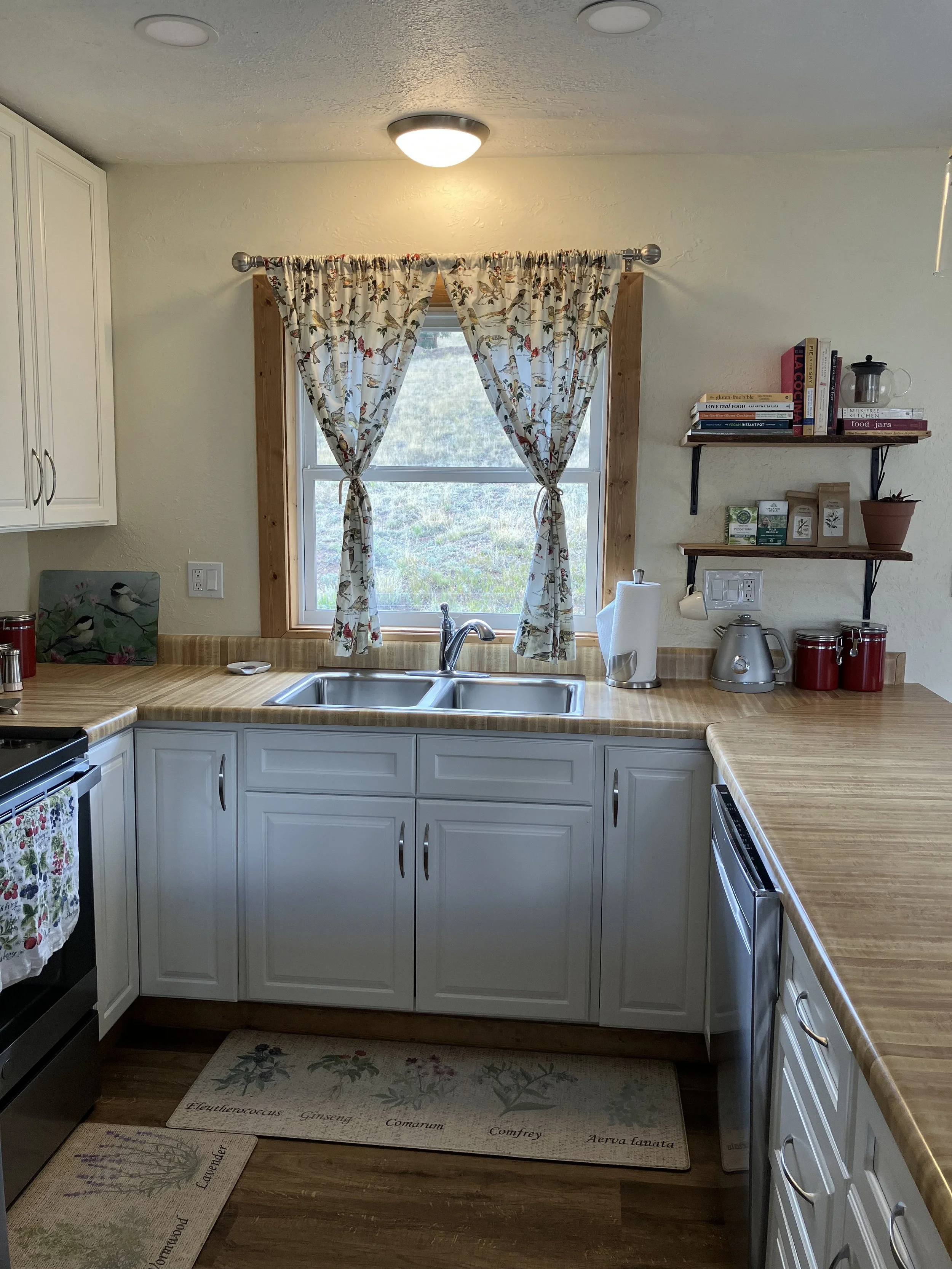 Kitchen with white cabinets, a window with floral curtains, a double sink, and wooden countertops. There are shelves with books and kitchen items, and a stove with a floral towel hanging on its handle.
