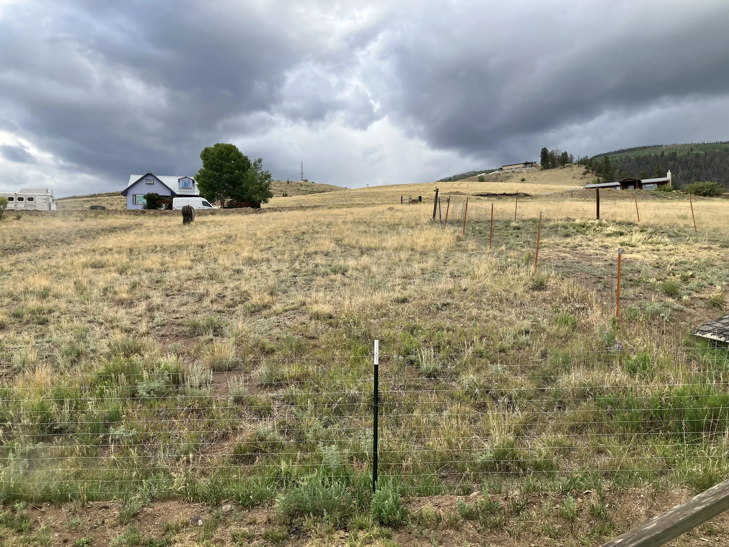 Open field with a fence in the foreground, a blue house with a white van and trees nearby on the left, and rolling hills with scattered houses under dark cloudy skies.