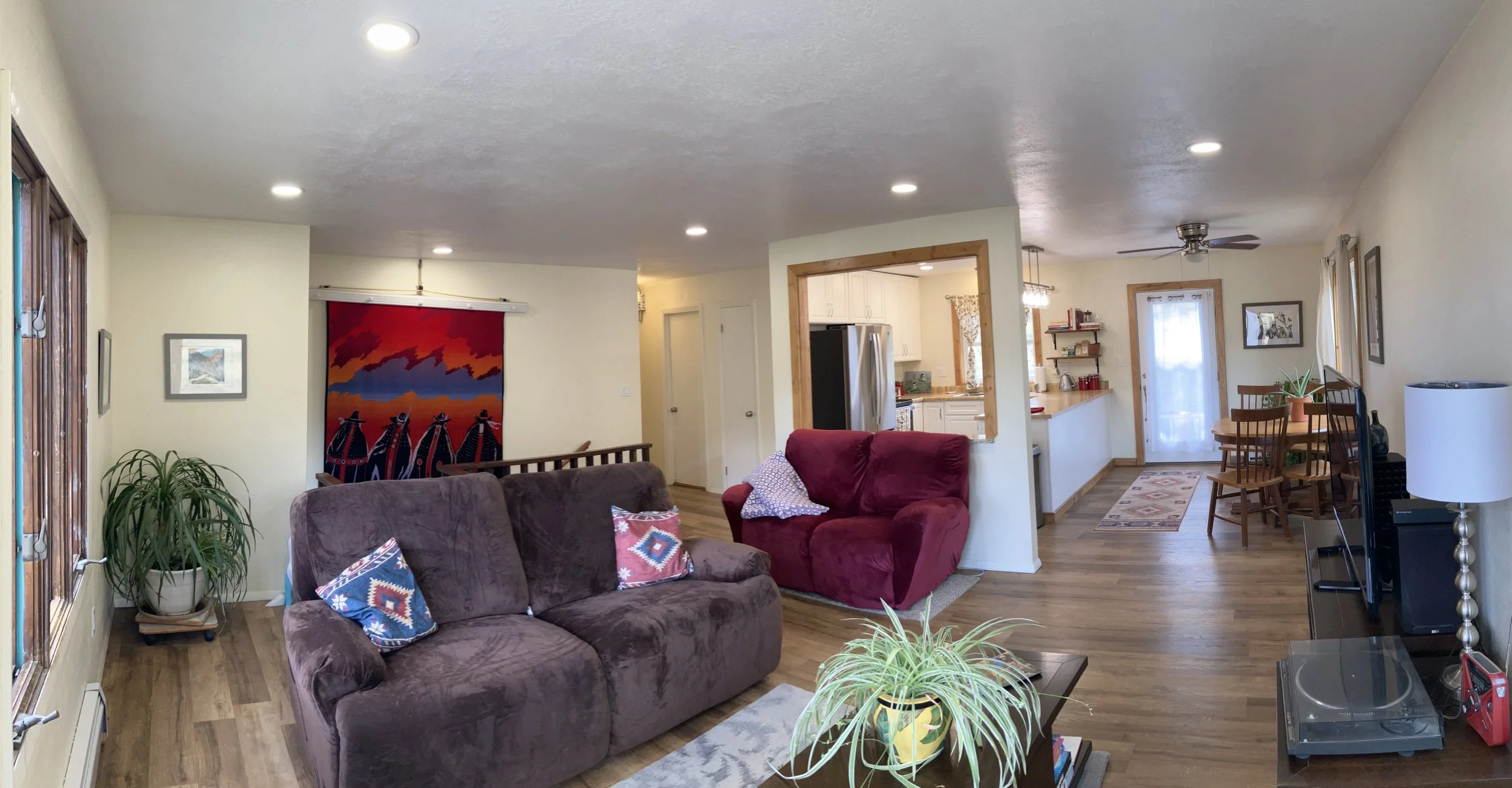 Living room with brown and red sofas, decorative pillows, houseplants, hardwood floors, artwork on walls, and open view to dining area and kitchen.
