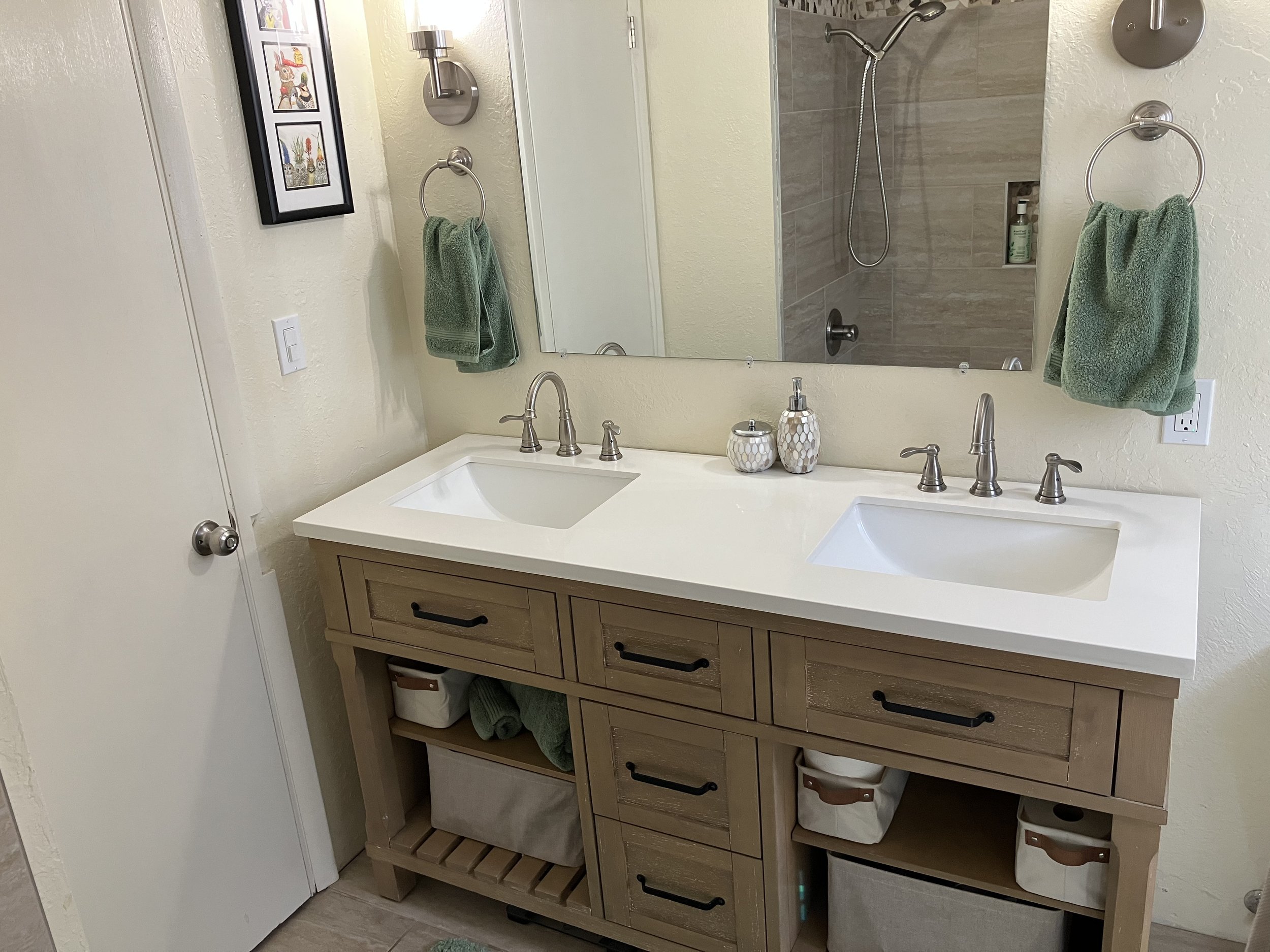 Bathroom with dual sinks, wooden vanity, large mirror, green towels, framed artwork, and a shower with a tiled wall, showerhead, and toiletries.