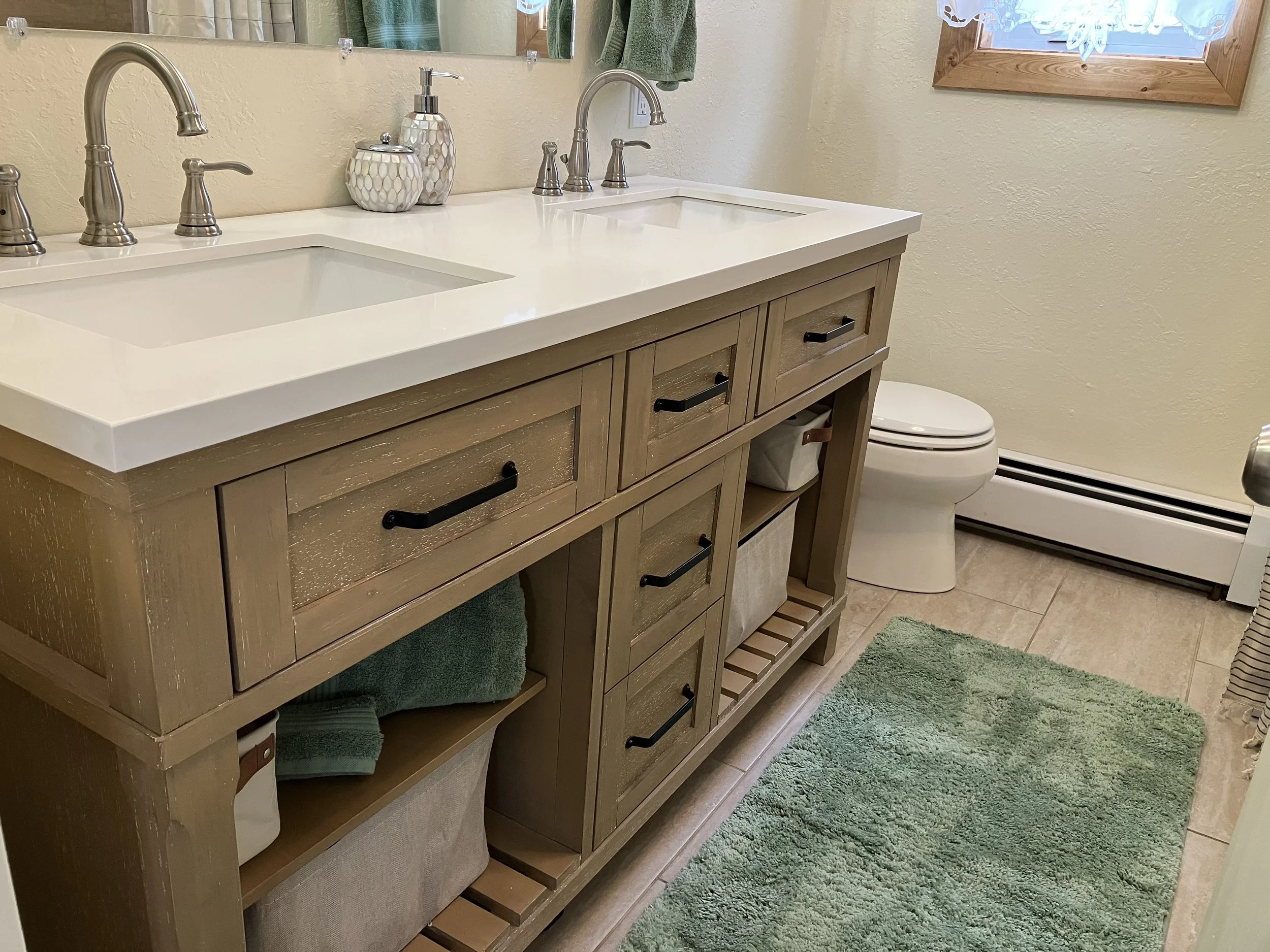 Bathroom vanity with two sinks, ceramic soap dispenser, towel, and decorative items, next to a toilet and a green bath mat.