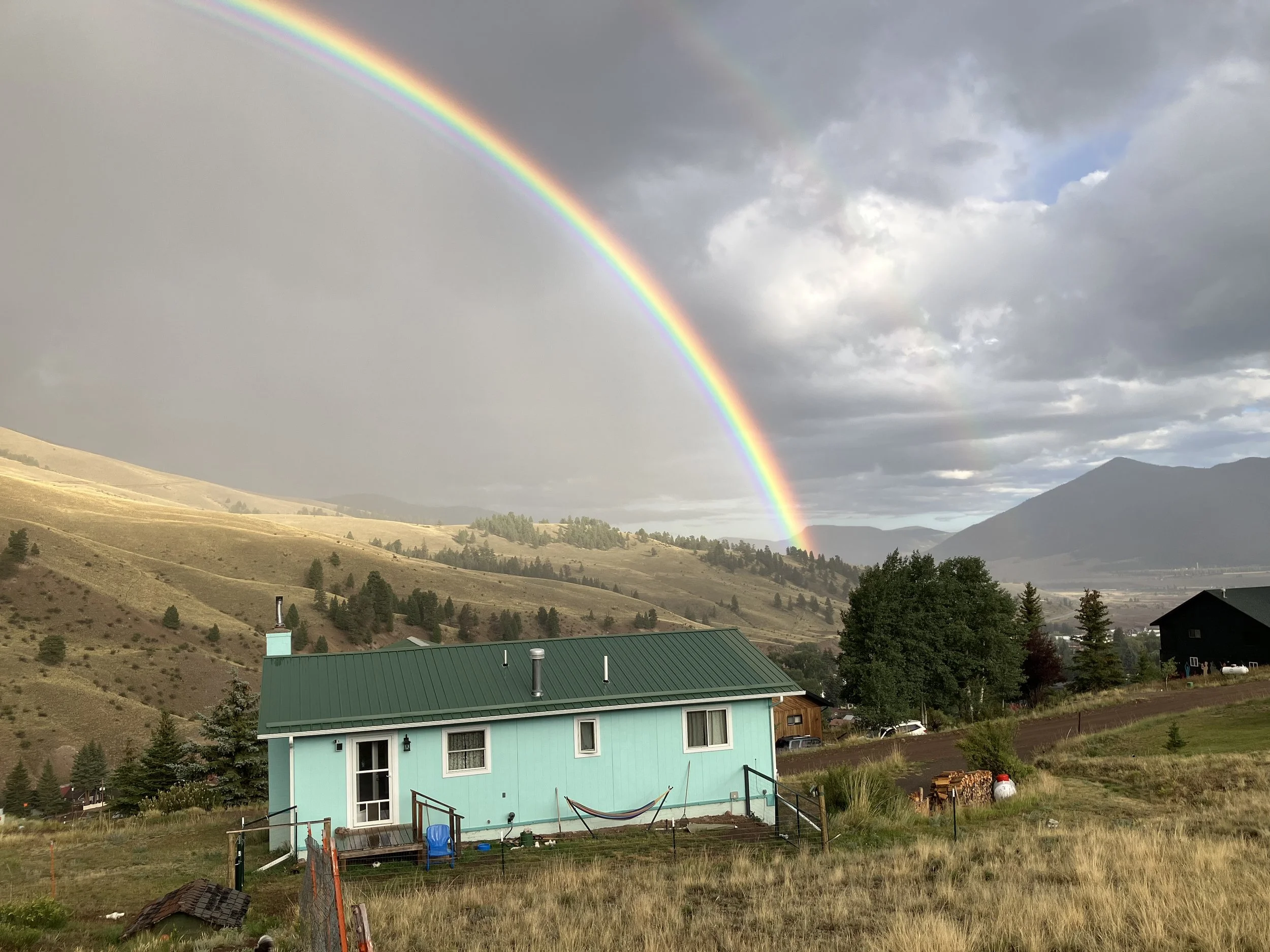 Scenic landscape with a vibrant rainbow arching over a green house with a metal roof in a rural area, surrounded by hills and trees, under a cloudy sky with patches of sunlight.