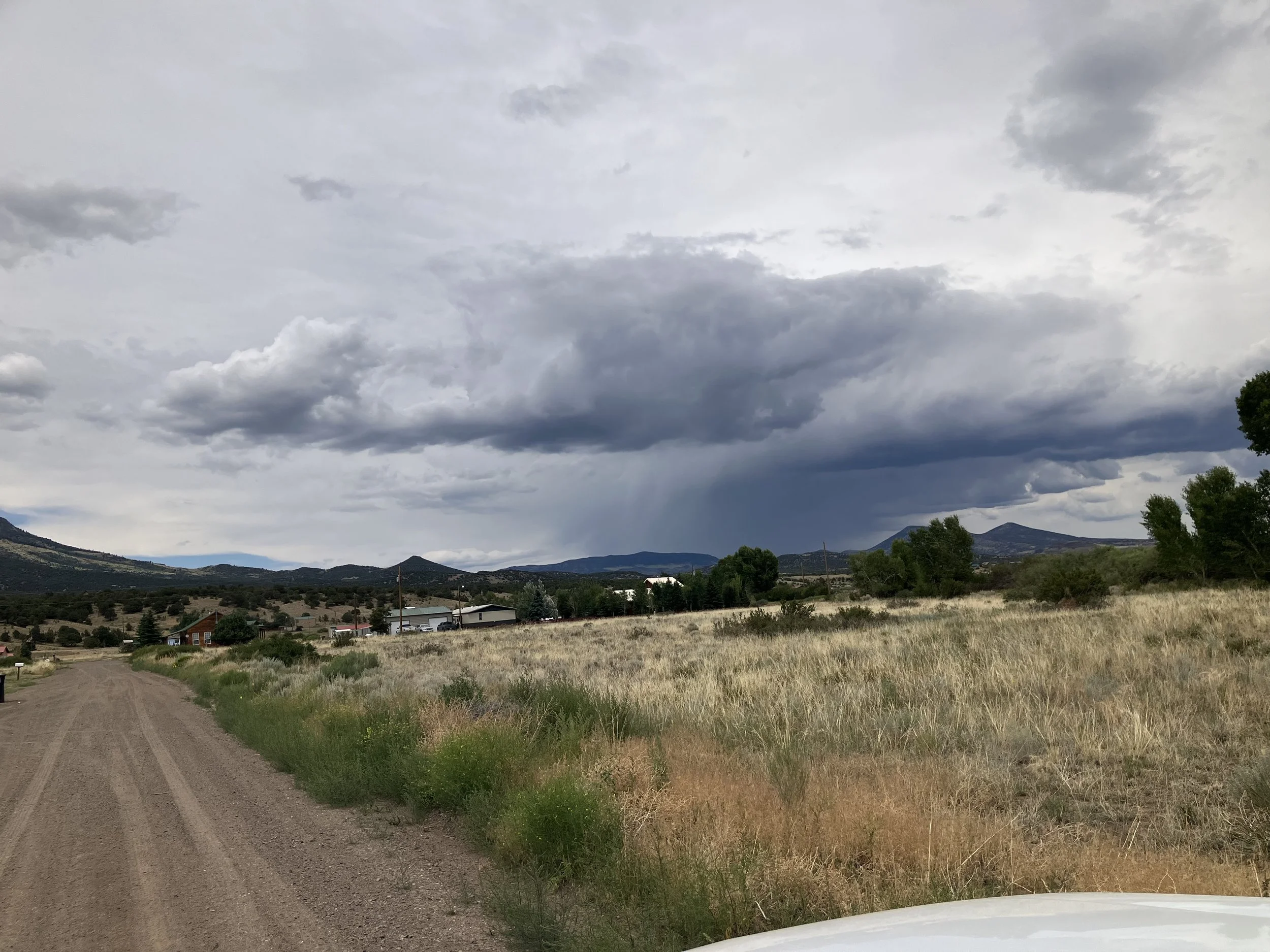 A rural landscape with a dirt road on the left, dry grassy fields, scattered trees, and mountains in the distance under a cloudy sky with dark storm clouds.