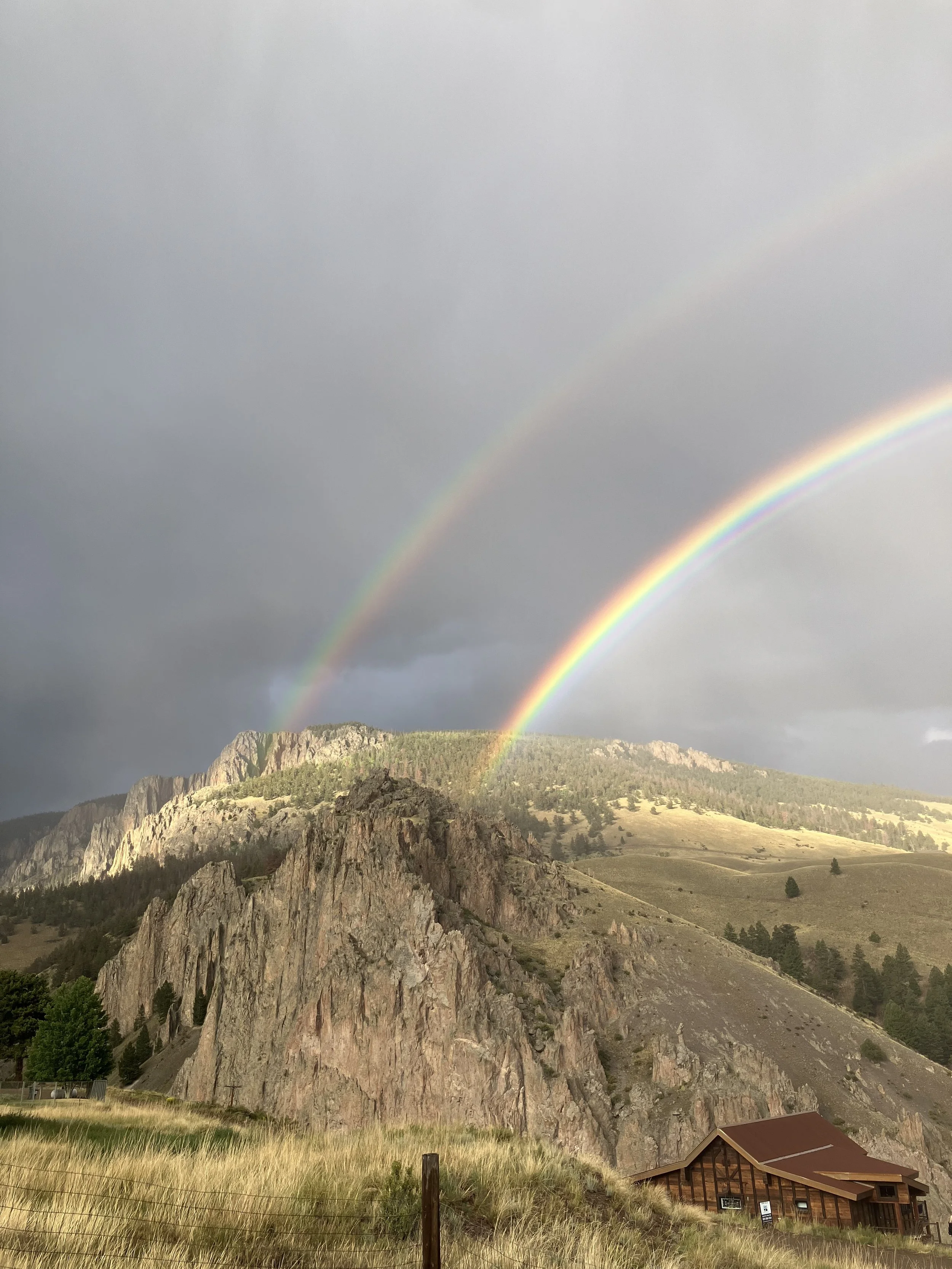 A landscape with rocky mountains, grassy fields, trees, and a wooden house. Two rainbows arc across a cloudy sky.