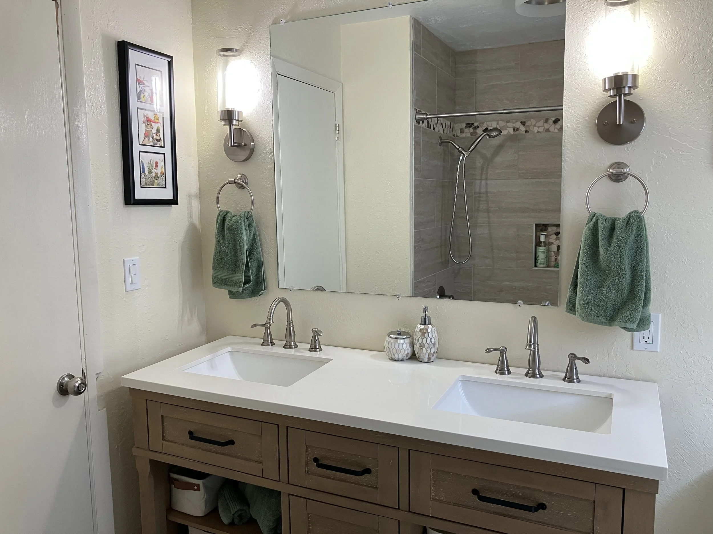 A bathroom vanity with two sinks, a large mirror, green towels, wall-mounted lights, a framed picture, and a view of a shower area with tiled walls.