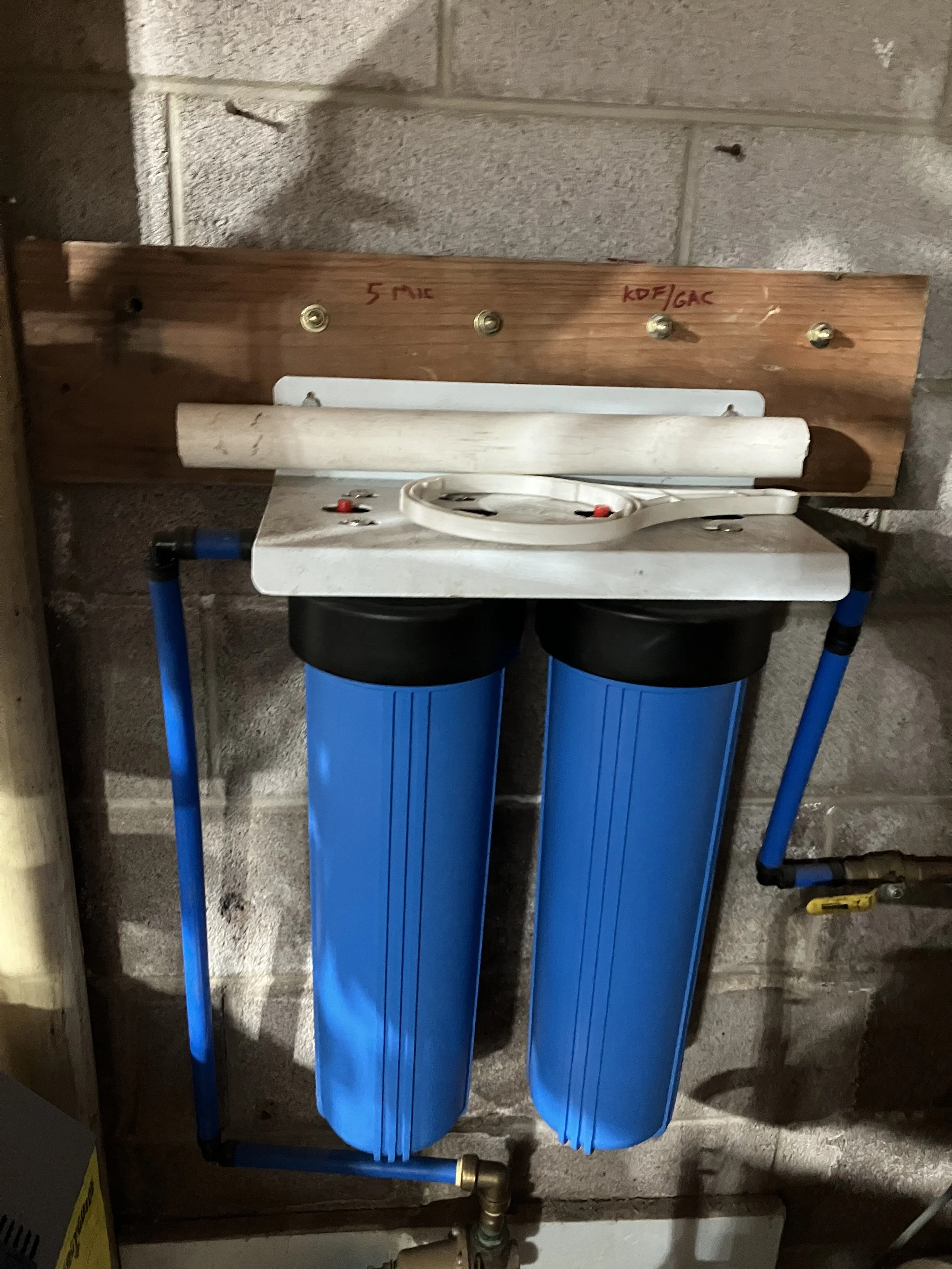 Water filtration system with two large blue filters mounted on a wall, connected by blue piping, with a wooden board above it labeled with red marker.