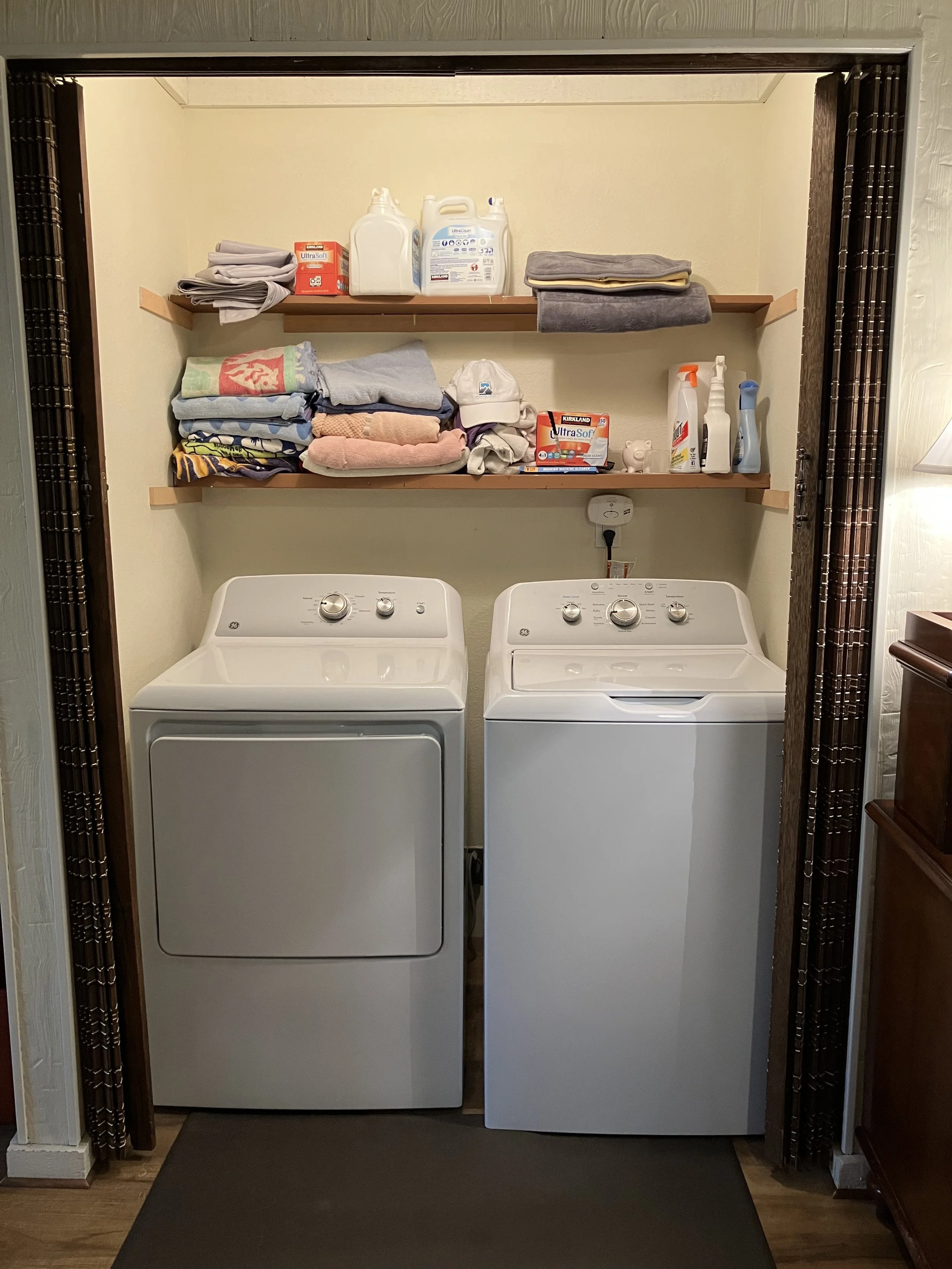 Laundry room with top-loading washing machine on the right and front-loading dryer on the left, with shelves above containing laundry supplies and folded towels.