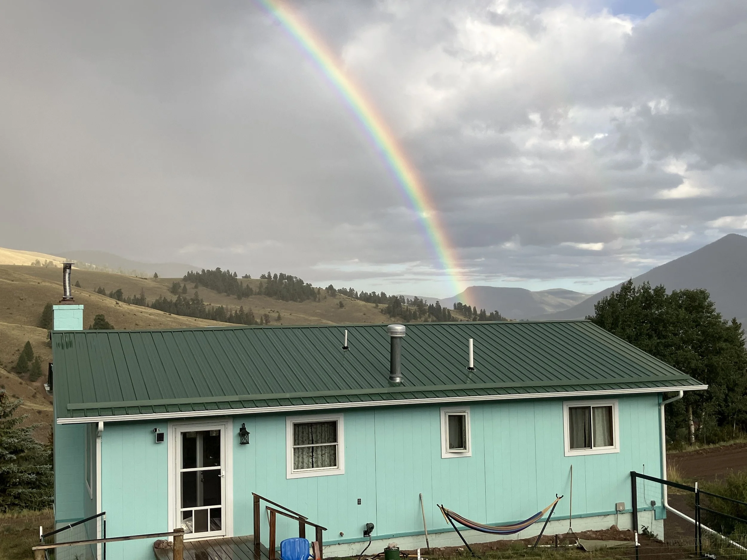 A turquoise house with a metal roof and a hammock in the front yard. A rainbow appears in the sky behind the house, with mountain hills in the background.