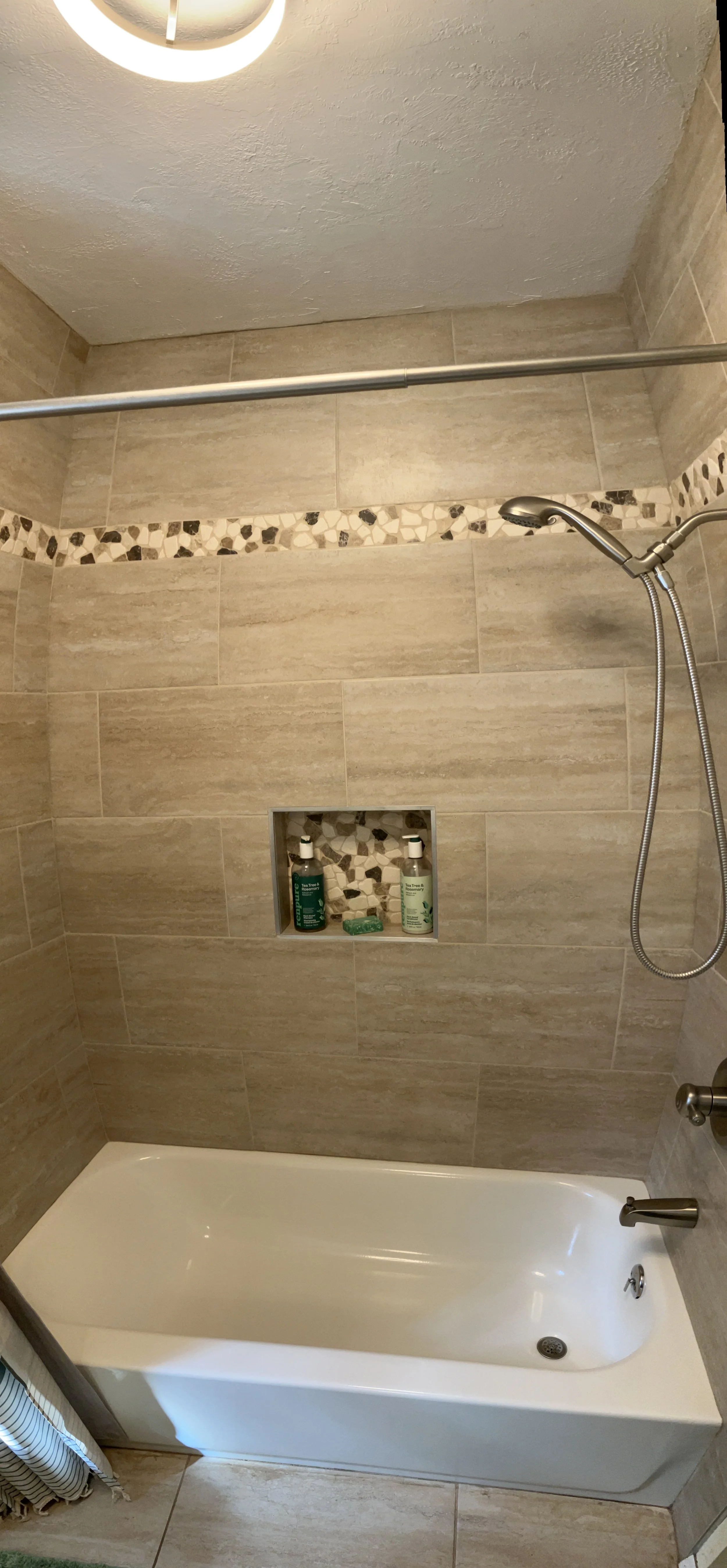 Bathroom shower with beige tiles, a built-in niche containing bottles of shower products, a bathtub, and a showerhead with a flexible hose.