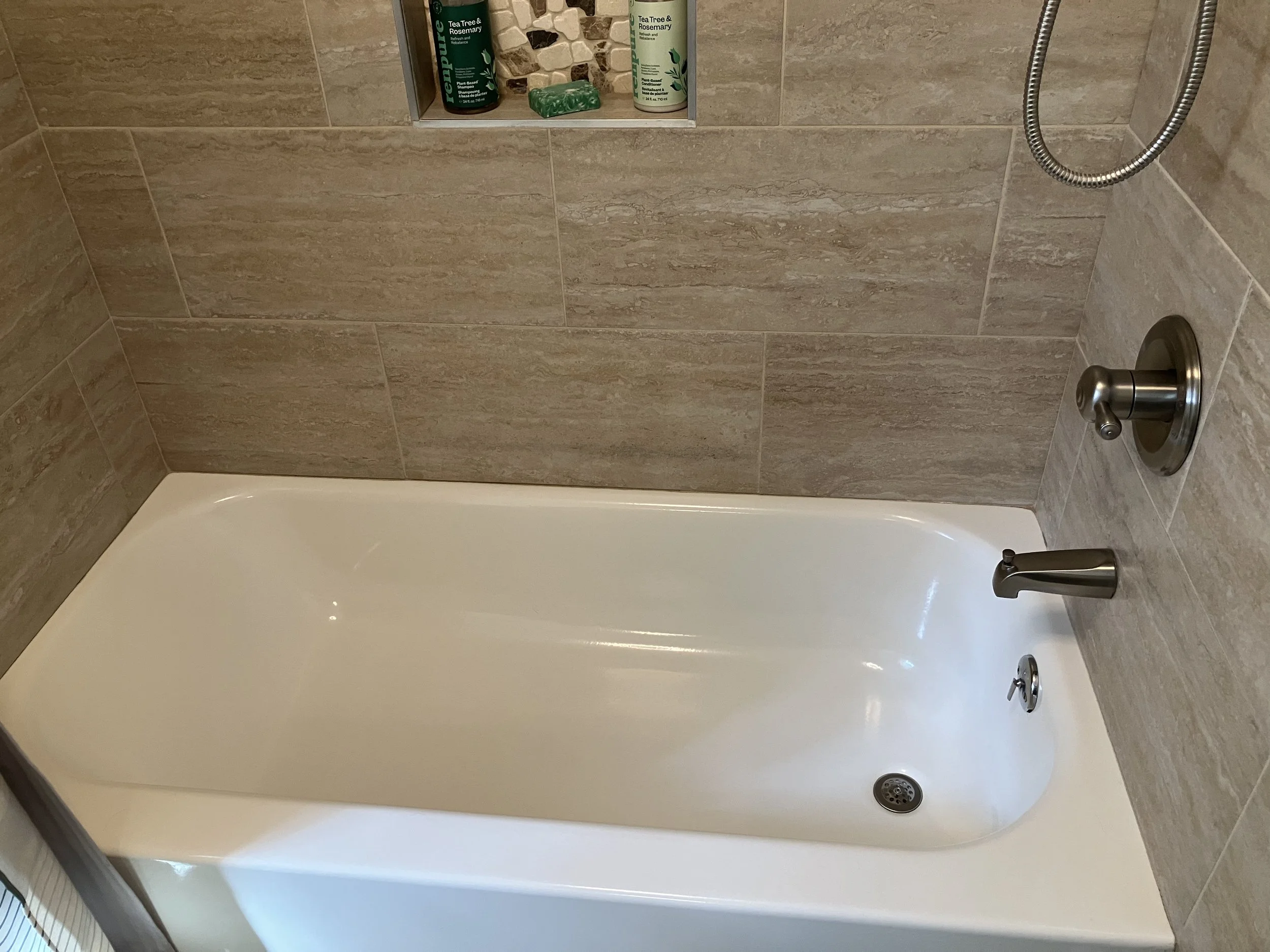 Empty white bathtub in a bathroom with beige tile walls, a brushed nickel faucet, and a built-in wall niche with toiletries including bottles of tea tree and rosemary shampoo and body wash.