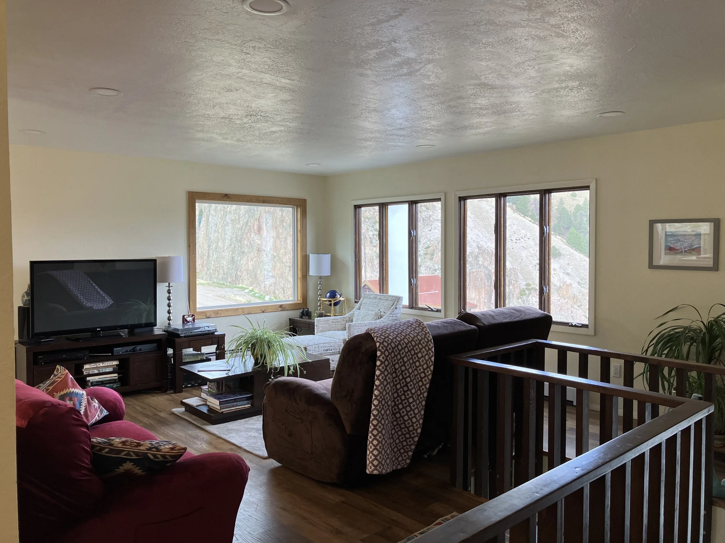 Living room with multiple large windows, a television on a stand, various seating including a red sofa, a brown recliner, and a white armchair, wooden flooring, plants, and framed pictures on the wall.