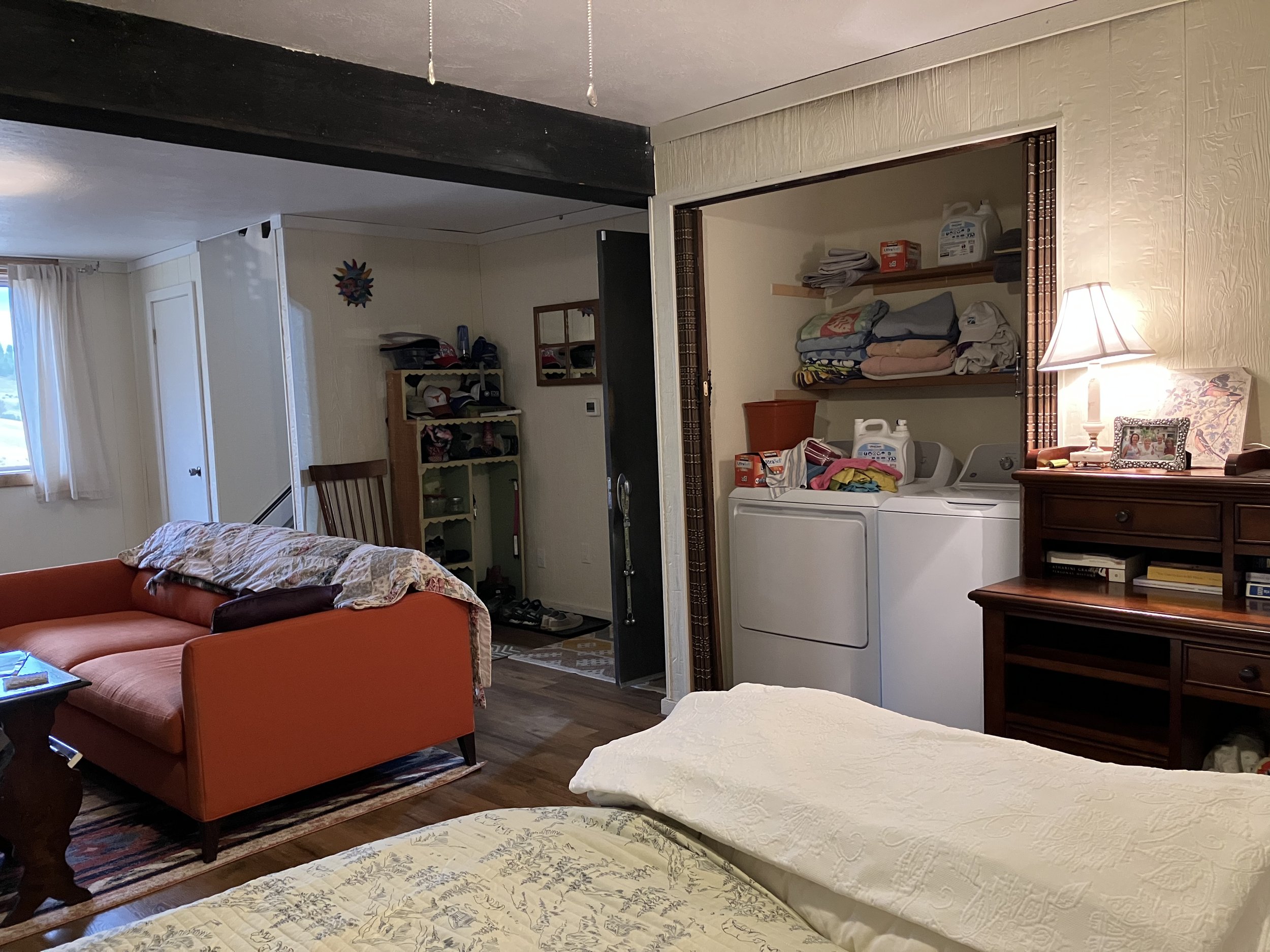 A cozy living room with a bed in the foreground, an orange sofa with a blanket, a window with curtains, a laundry nook with a washing machine and shelves, and a wooden dresser with a lamp and decorative items.